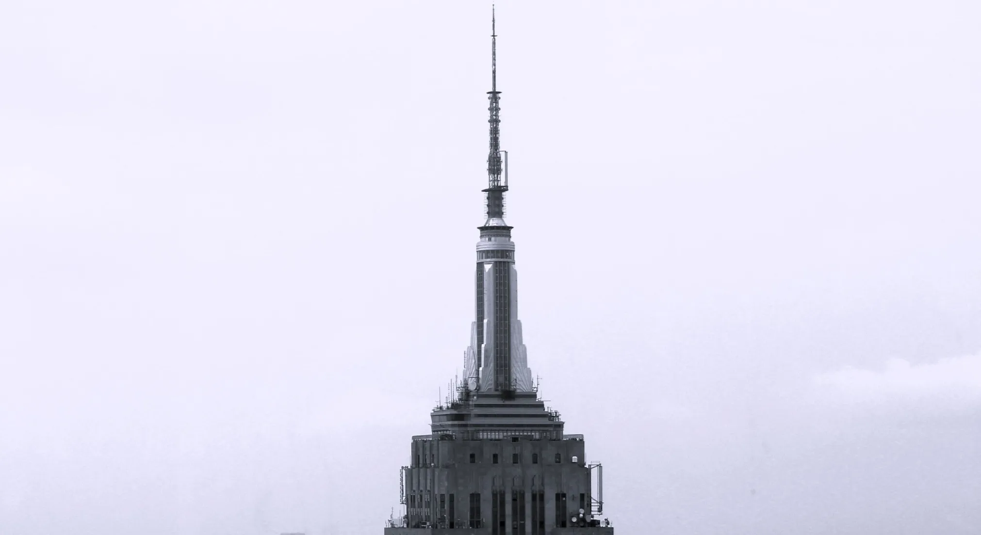 A zoomed-in black and white shot of a tall, thin skyscraper with a prominent spire against a pale sky. This is a landmark.