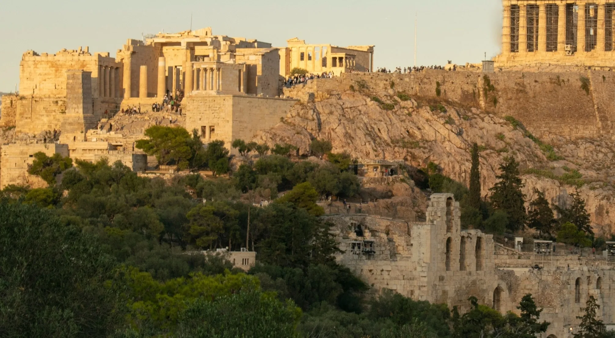 A zoomed-in image of ancient landmarks: the Acropolis of Athens and the Parthenon in golden light, with trees and ruins below.
