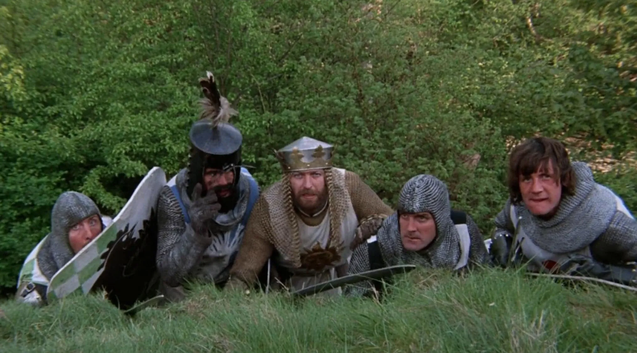 Five knights from Monty Python comedy group peering over a grassy knoll, dressed in medieval armor.