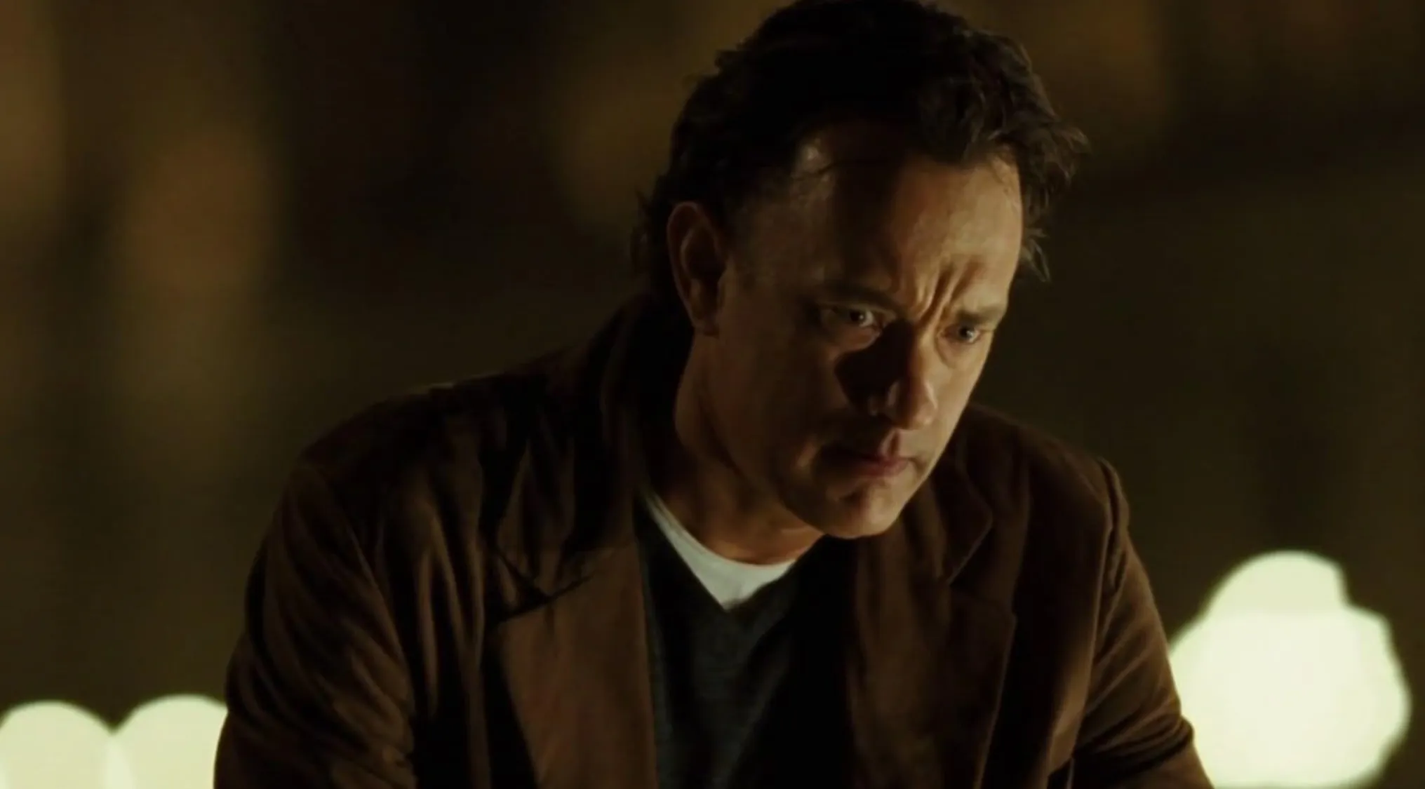 Close-up of Robert Langdon, played by Tom Hanks, looking serious in a dark setting, from a movie franchises.