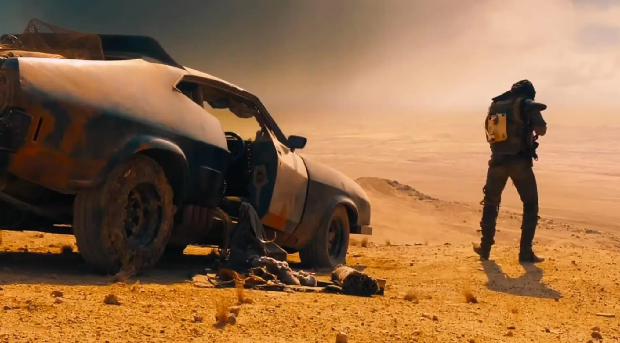 A lone figure surveys a desolate desert landscape beside a wrecked vehicle, reminiscent of the Mad Max film franchise.