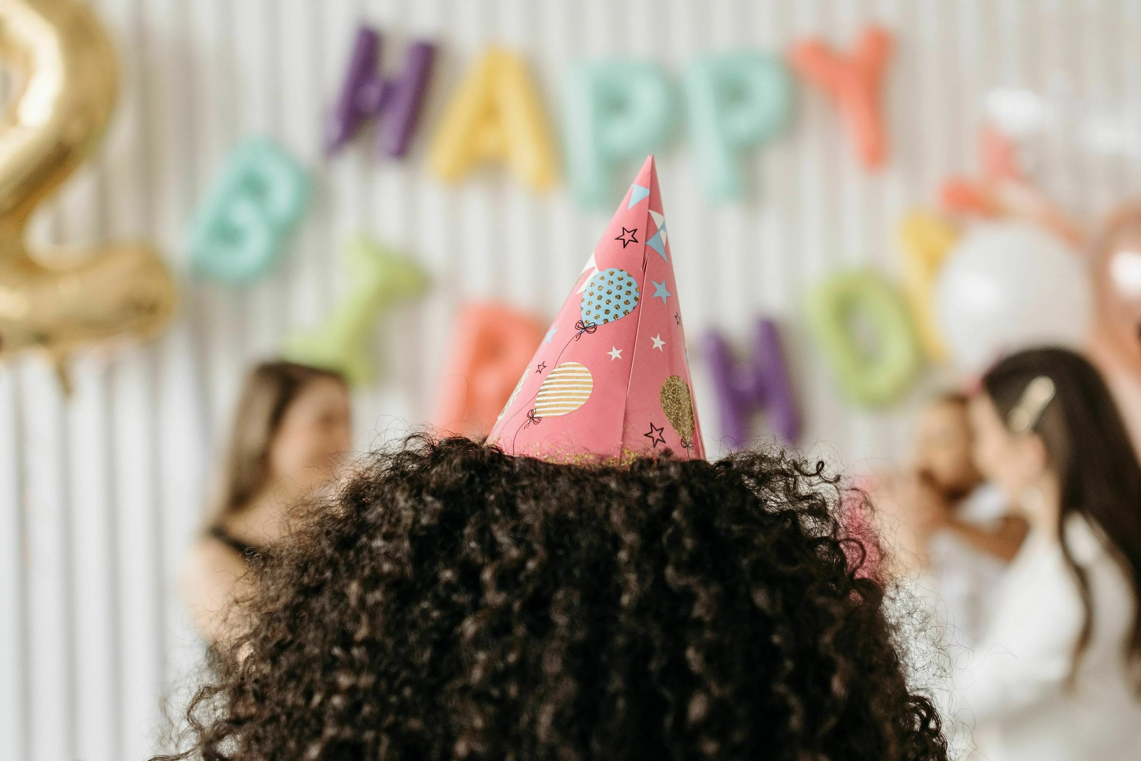 The back of a head with curly hair wearing a pink party hat at a birthday party. A mystery party guest at a fun party.