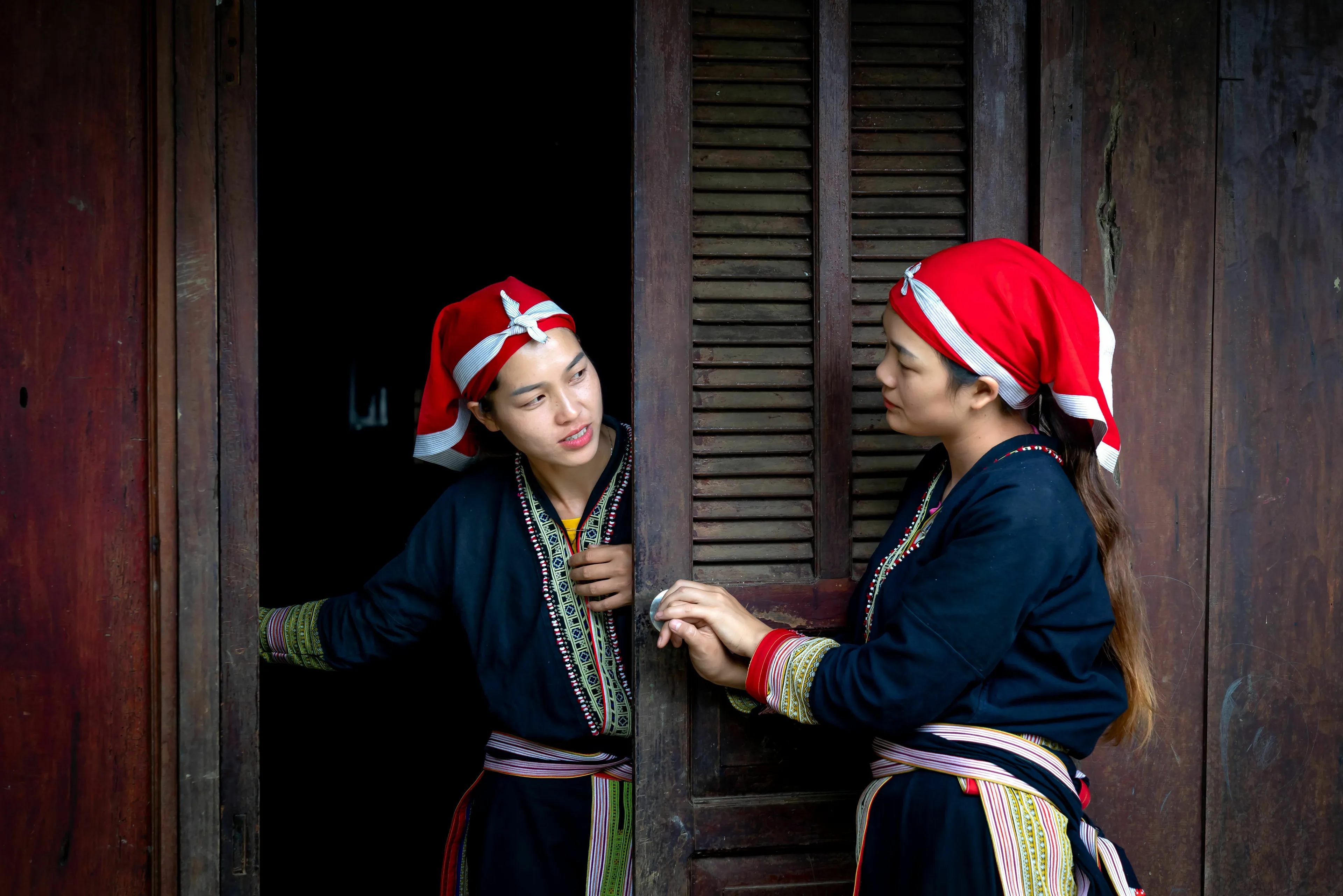 Two women in traditional attire by a wooden door, discussing what meal to have for a loved one.