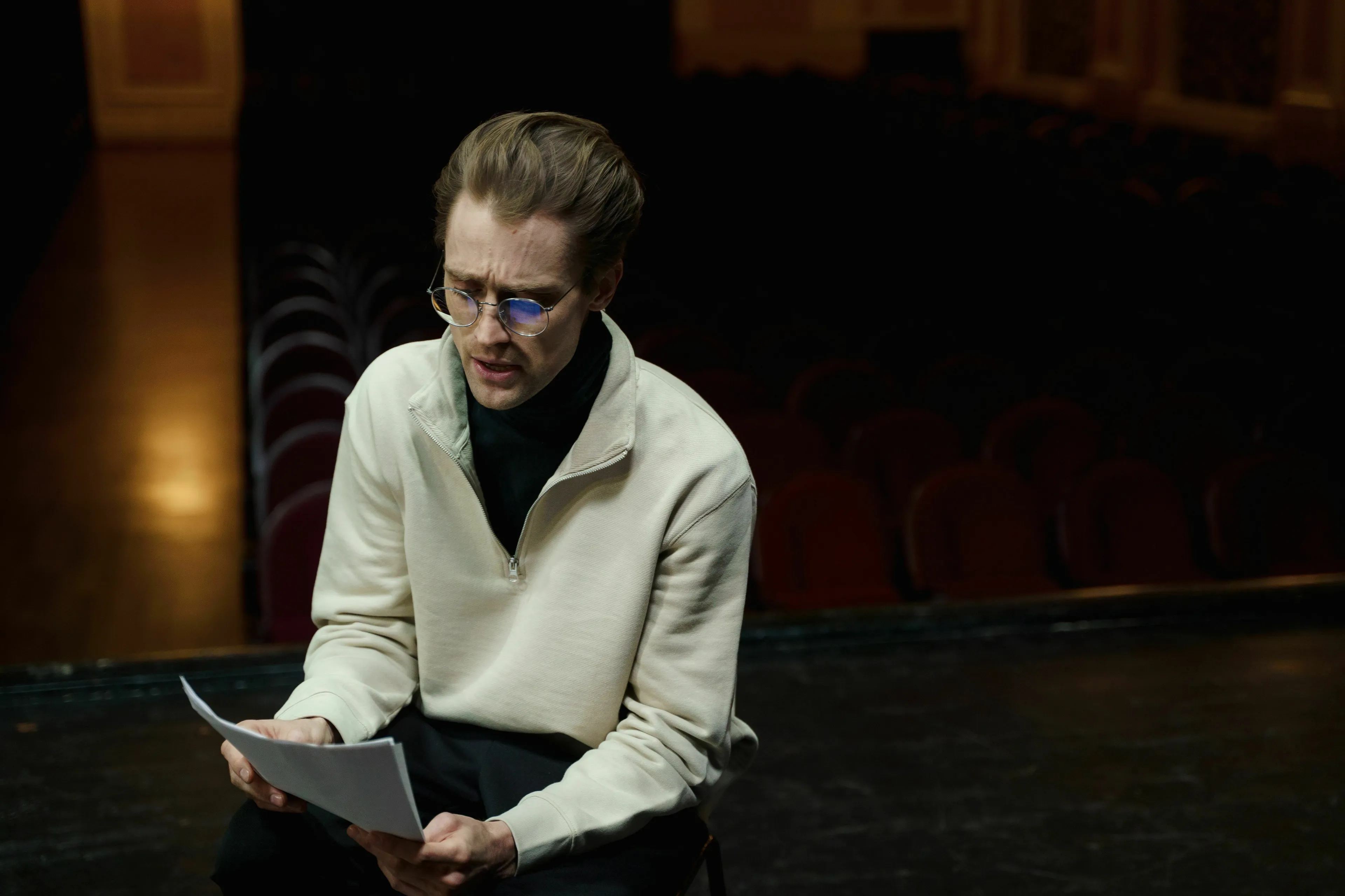 Movie villain rehearsing a monologue, sitting on a stage with papers in hand.