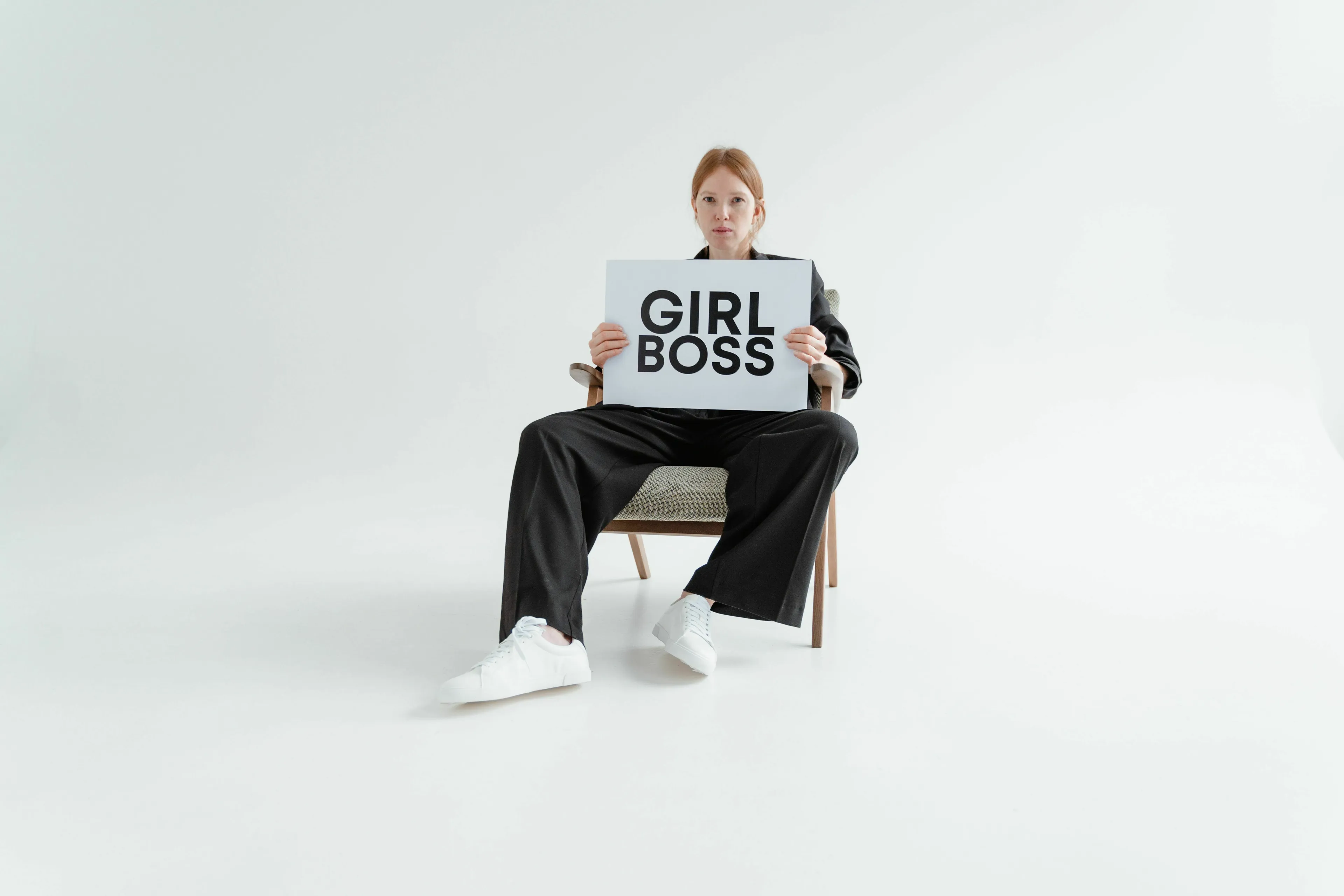 A woman in a black suit sits on a chair, holding a 'GIRL BOSS' sign, representing a movie villain's treatment of people.