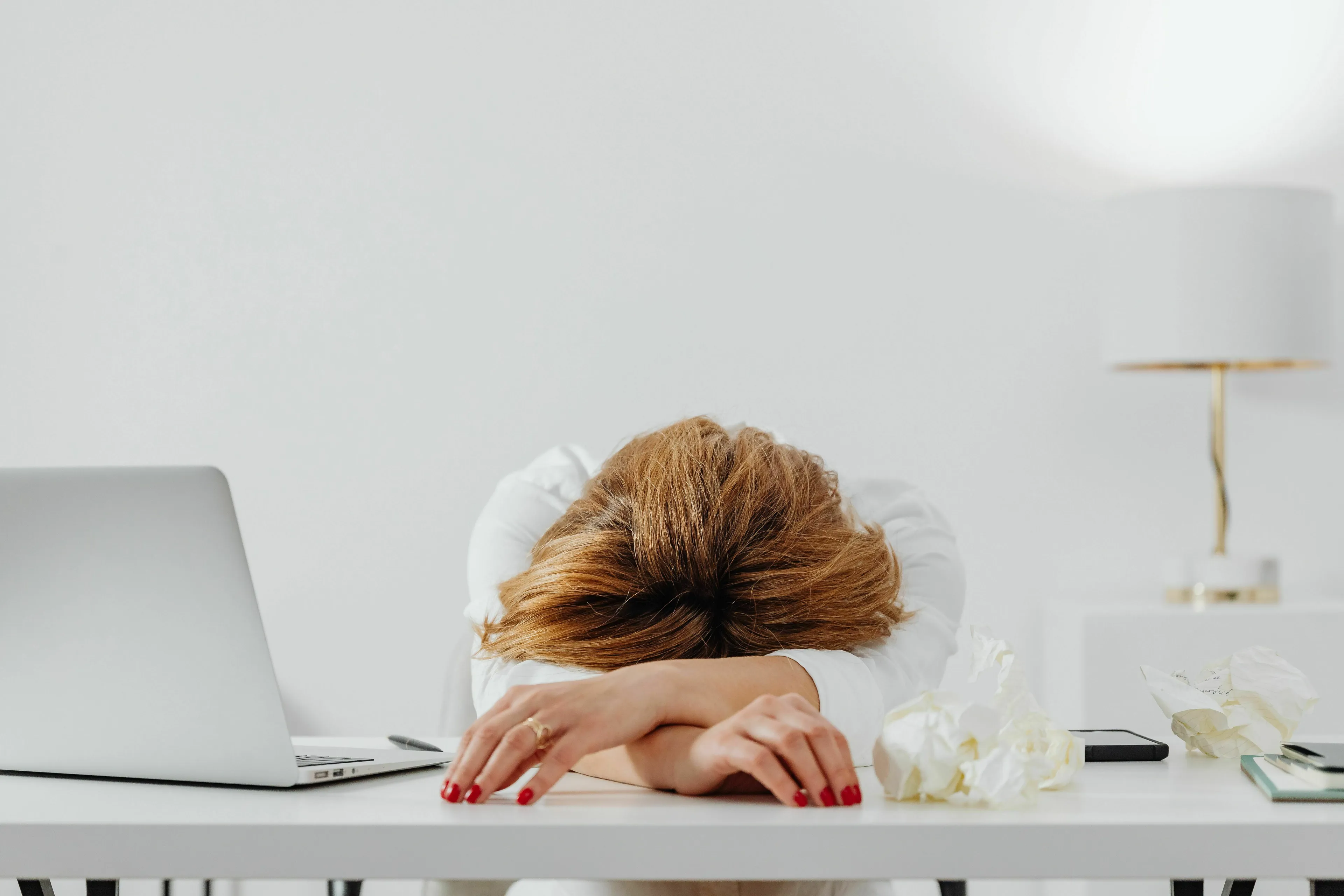 A person with red hair rests their head on a desk next to a laptop and crumpled tissues, depicting a week-ruining moment for a movie villain.