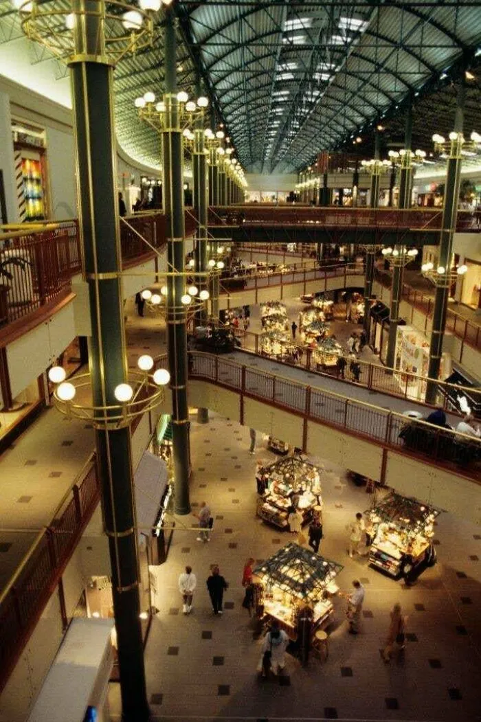 A sprawling mall interior from the 90s, featuring multiple levels and brightly lit kiosks, evoking nostalgia for 90s life.