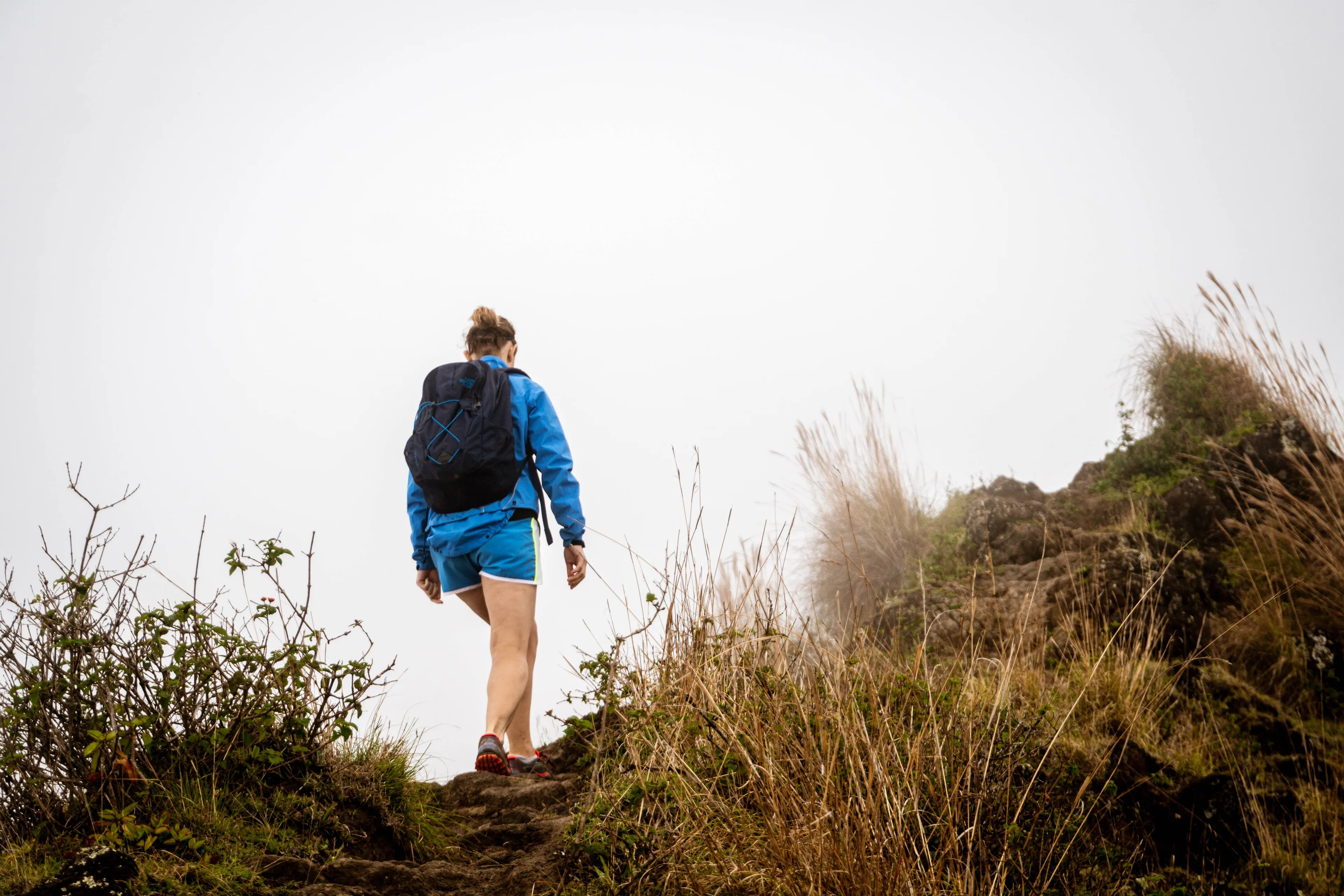 A person with a backpack taking a solo hike up a grassy, rocky trail on an overcast day.