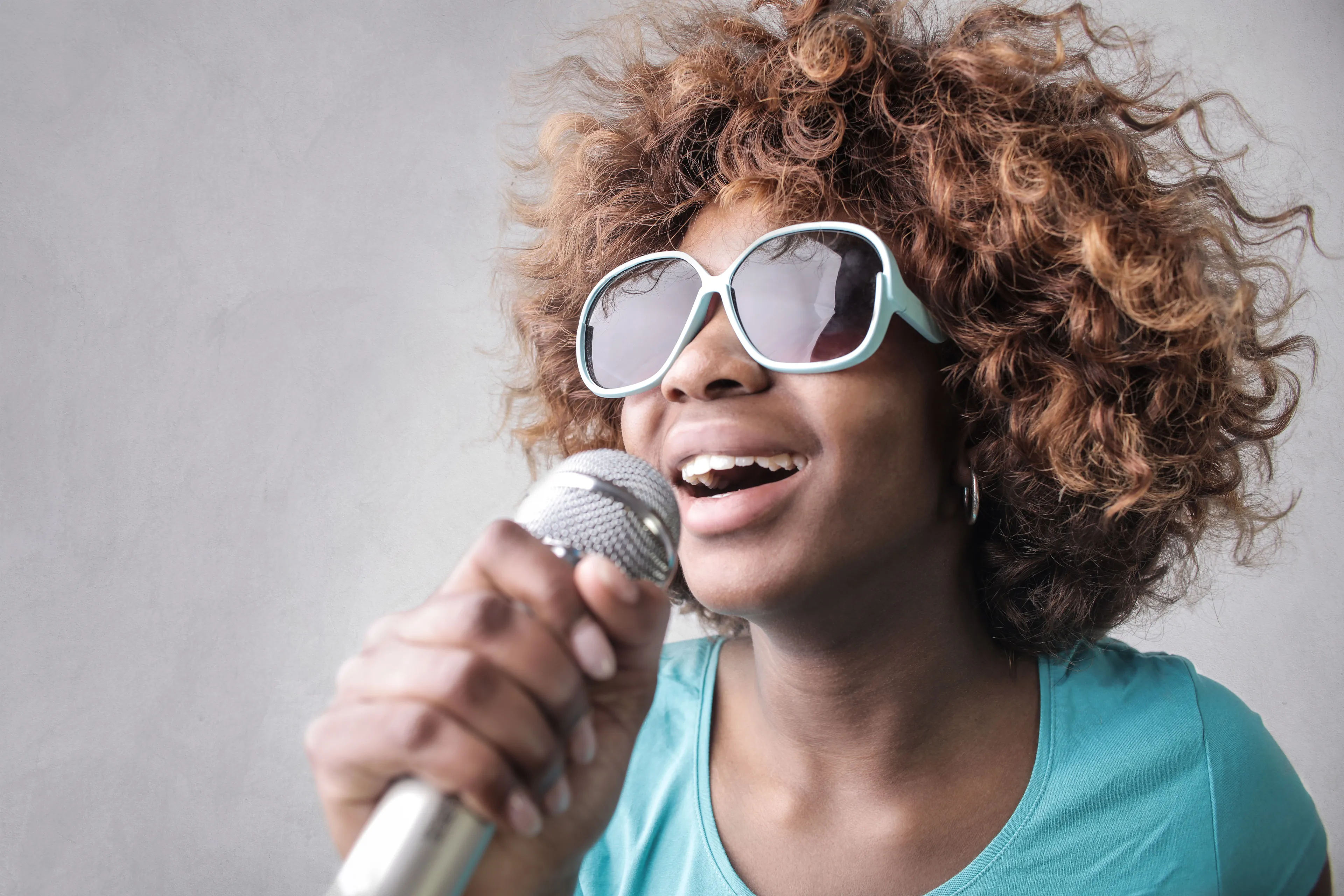 A young woman with curly hair wearing sunglasses, singing enthusiastically into a microphone at an open mic event.