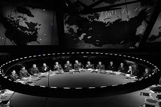 A black and white image of men in military uniforms seated around a large round table in the war room from Dr. Strangelove.