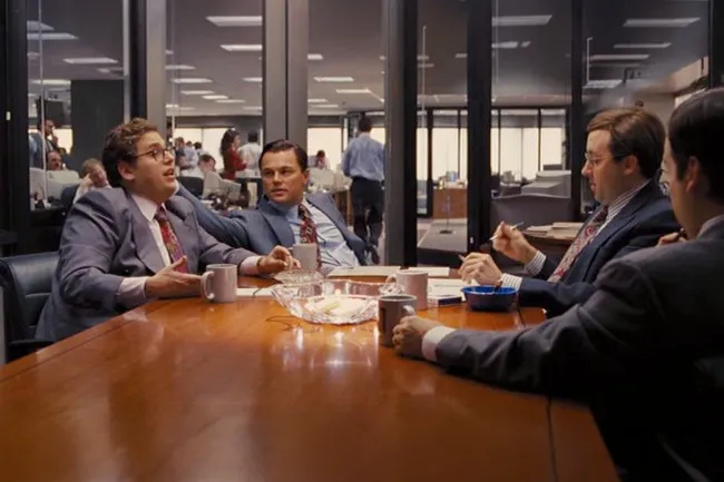 Men in suits sitting around a large wooden table in the boardroom from The Wolf Of Wall Street.