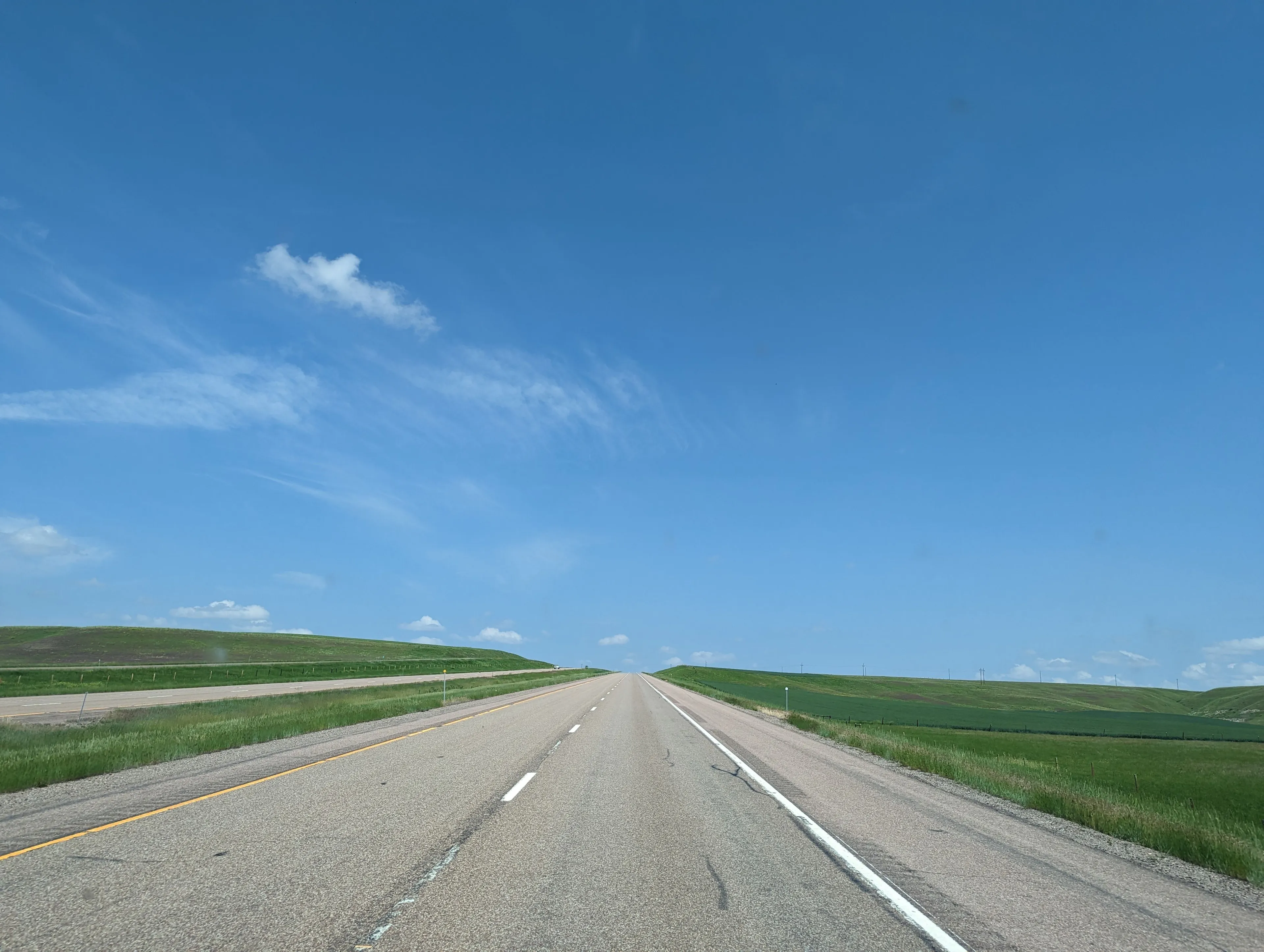 A long, clear road stretches through green fields under clear skies.
