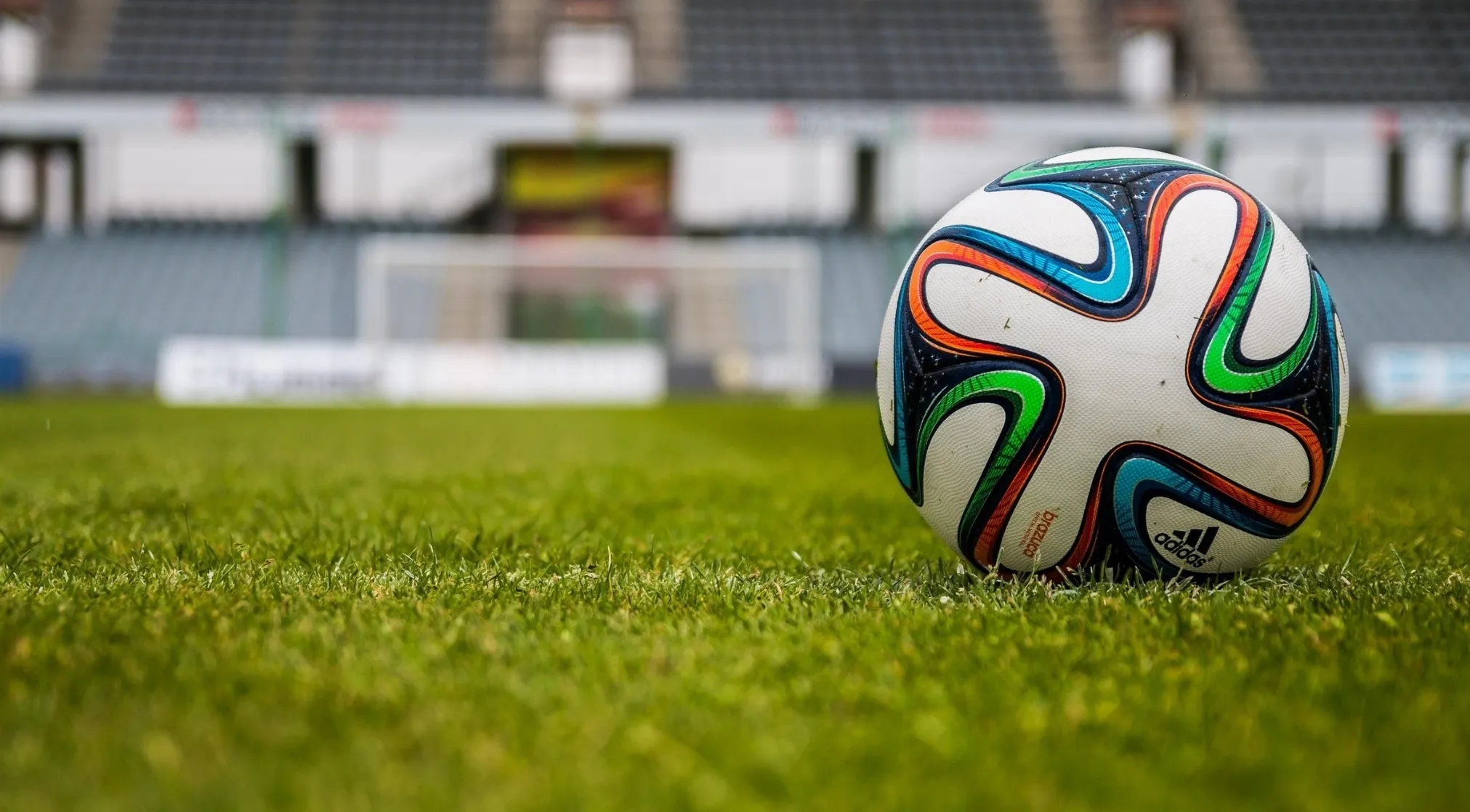 A FIFA World Cup soccer ball on lush green grass with stadium seats and a goal in the blurred background.