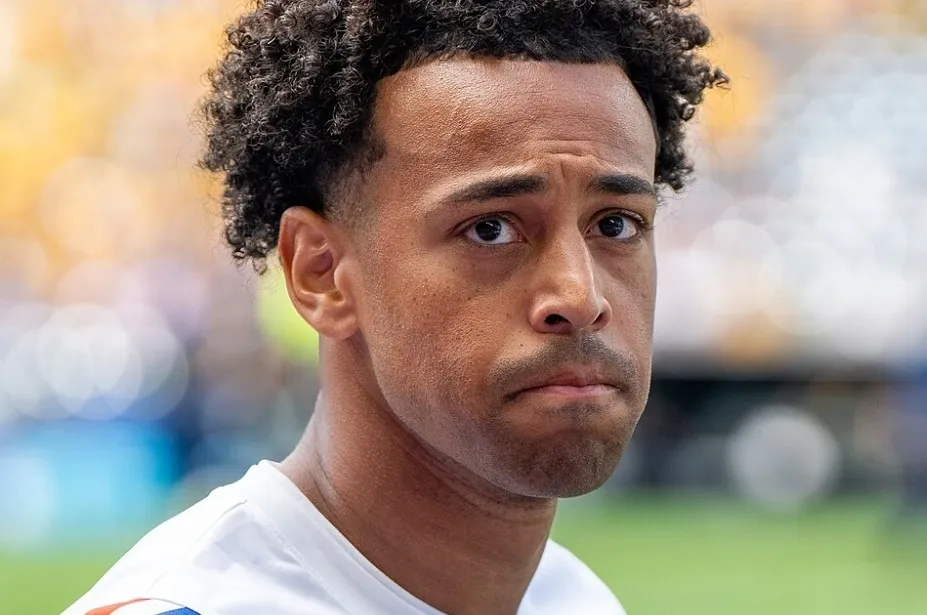 Close-up of Tyler Adams, nicknamed Captain America, with a serious expression, dark curly hair, and a light blurred background.