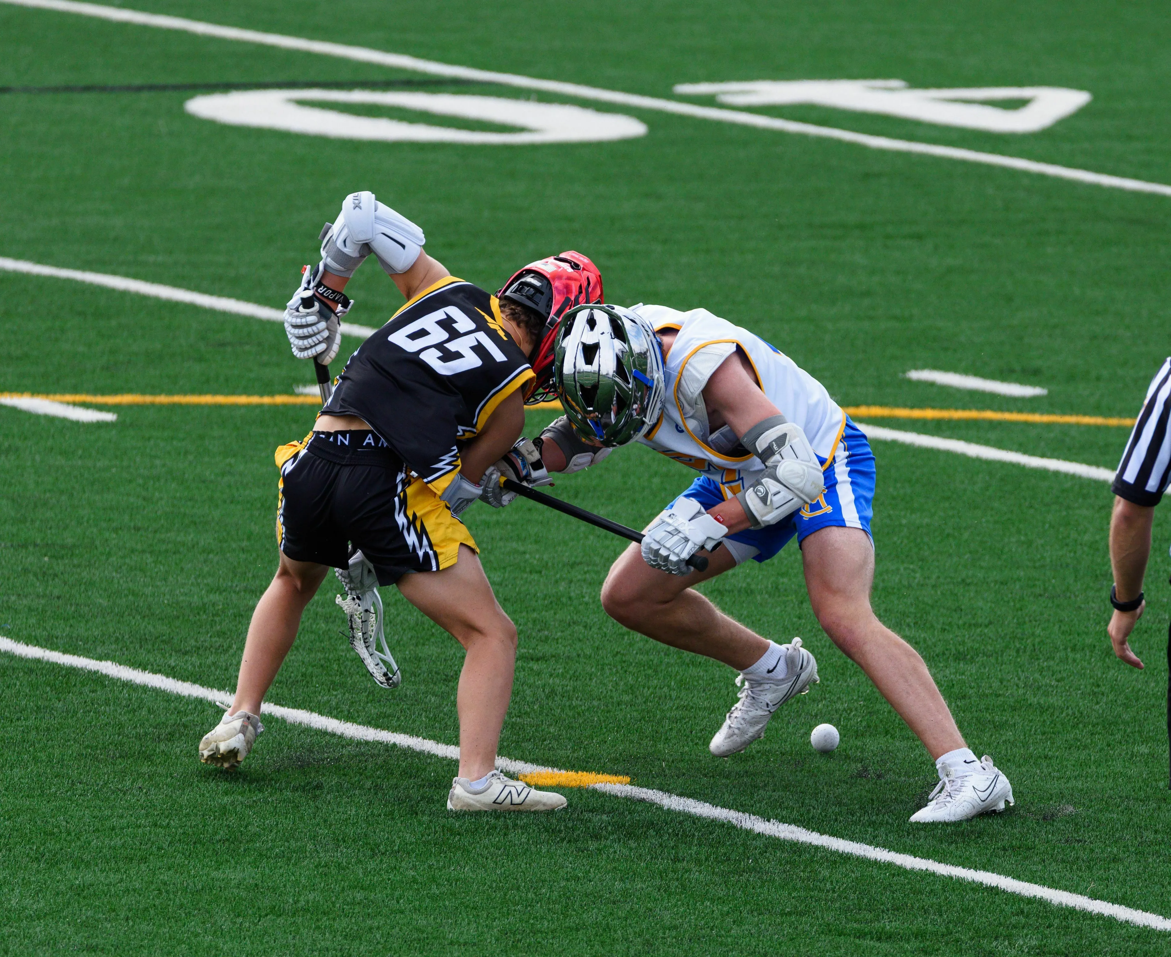 Two players compete for the ball during a lacrosse game on a green field.