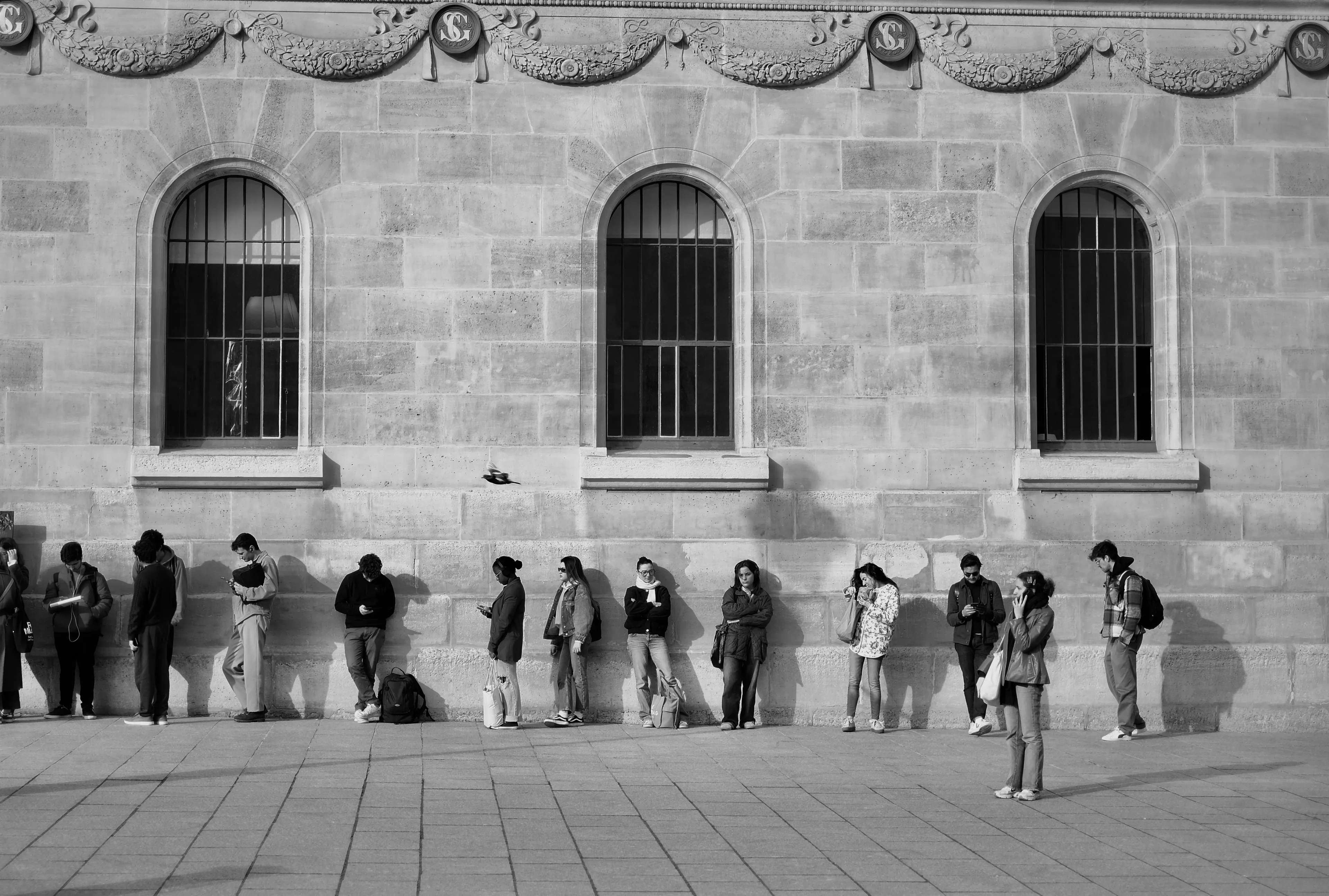 A black and white photo of people waiting in a slow-moving line outside a stone building, exploring relationship with rules.