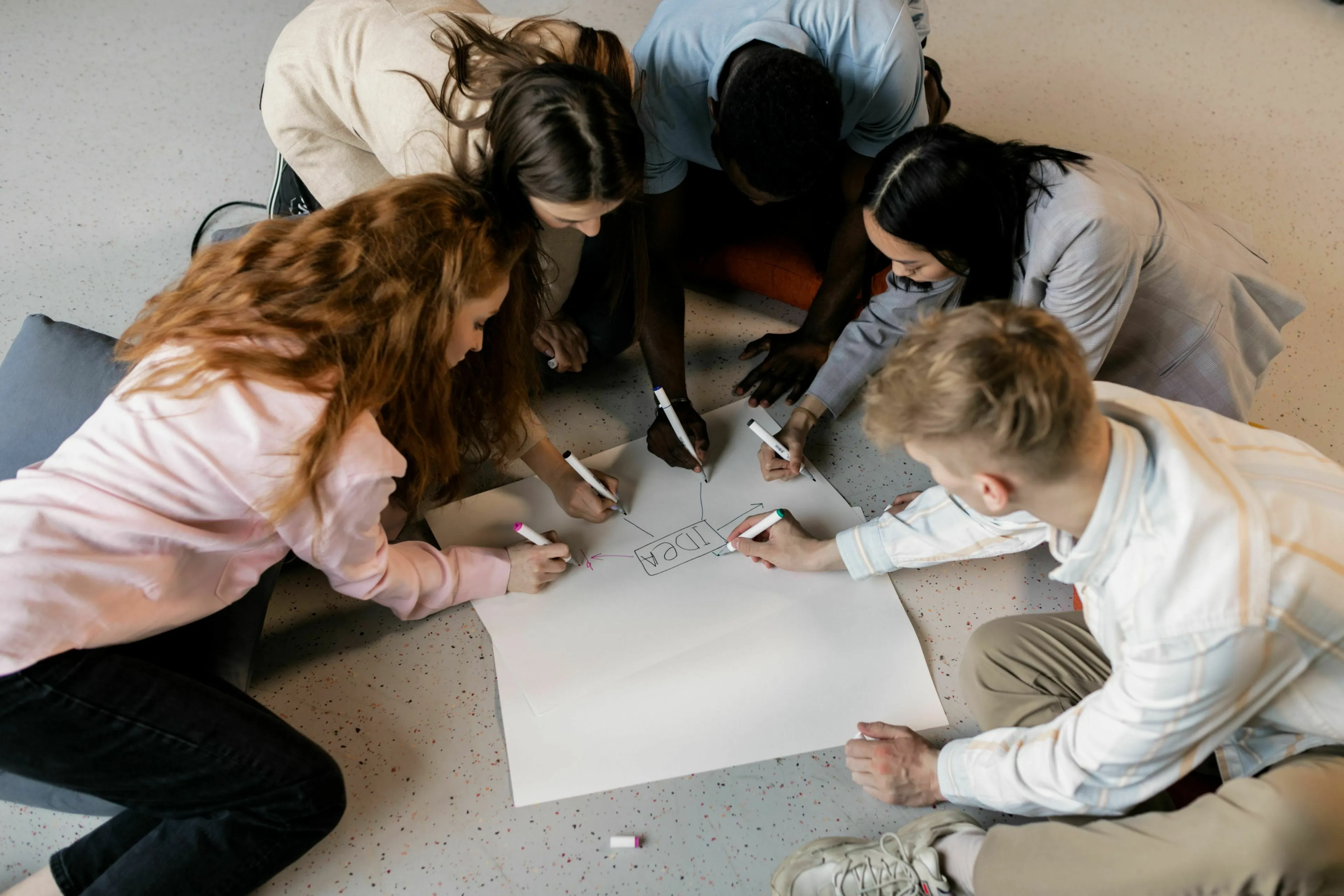 Diverse group project members collaborating on a large sheet of paper on the floor, brainstorming ideas.