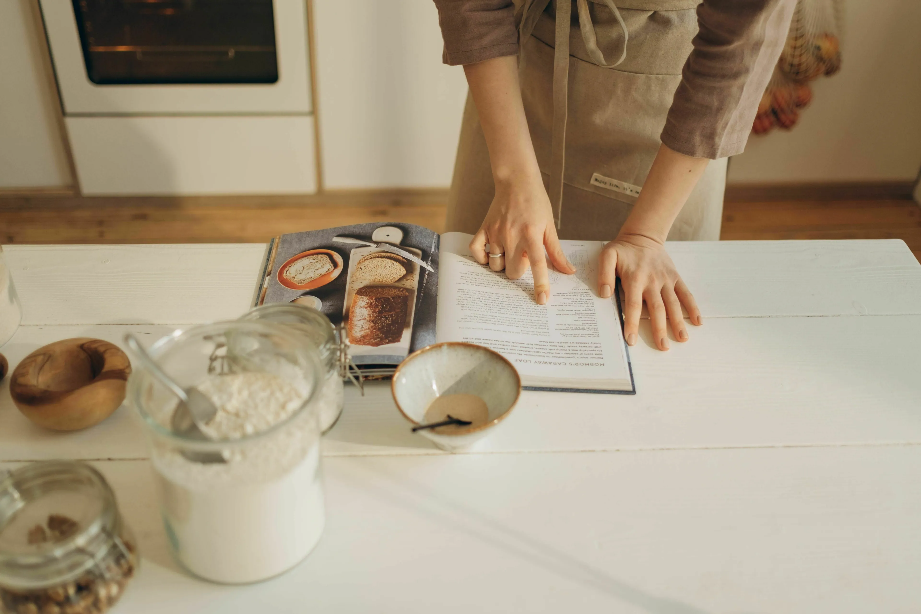 A person in an apron, hands pointing to a recipe in an open cookbook on a white kitchen counter.