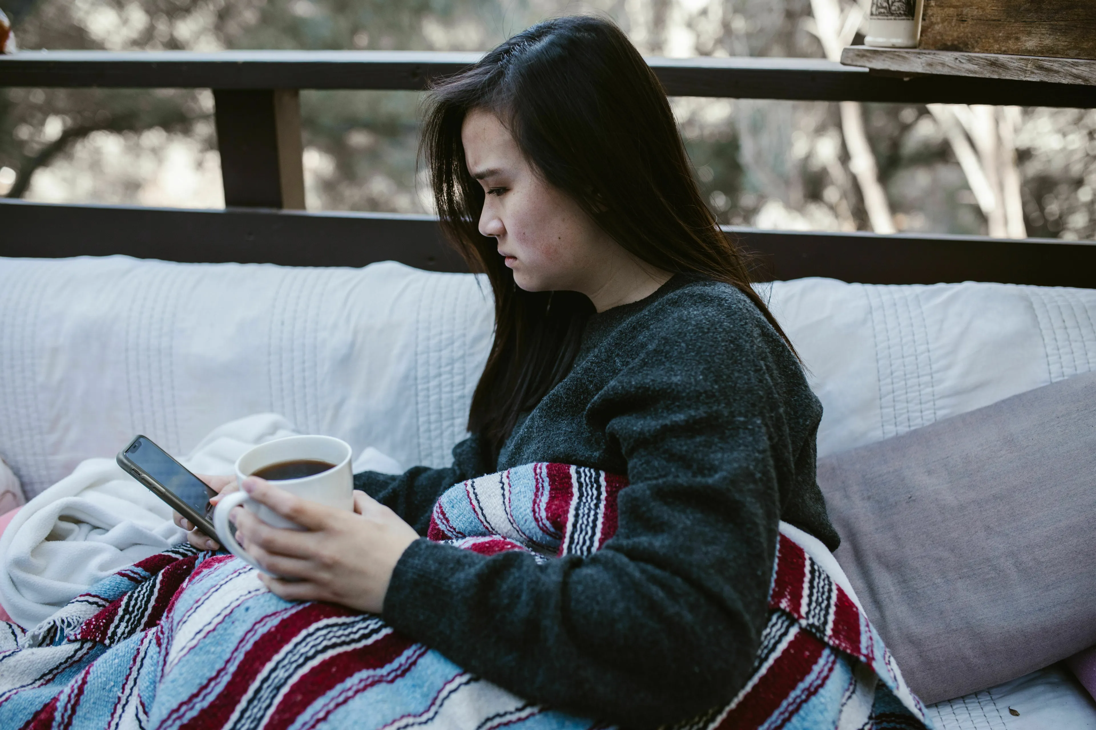 A young woman, covered in a blanket, looking intently at her phone while holding a mug, reacting to group chat rules.