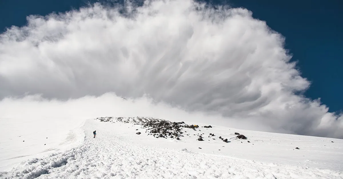 A lone person walks on a snow-covered mountain with a large cloud formation above, depicting an avalanche.