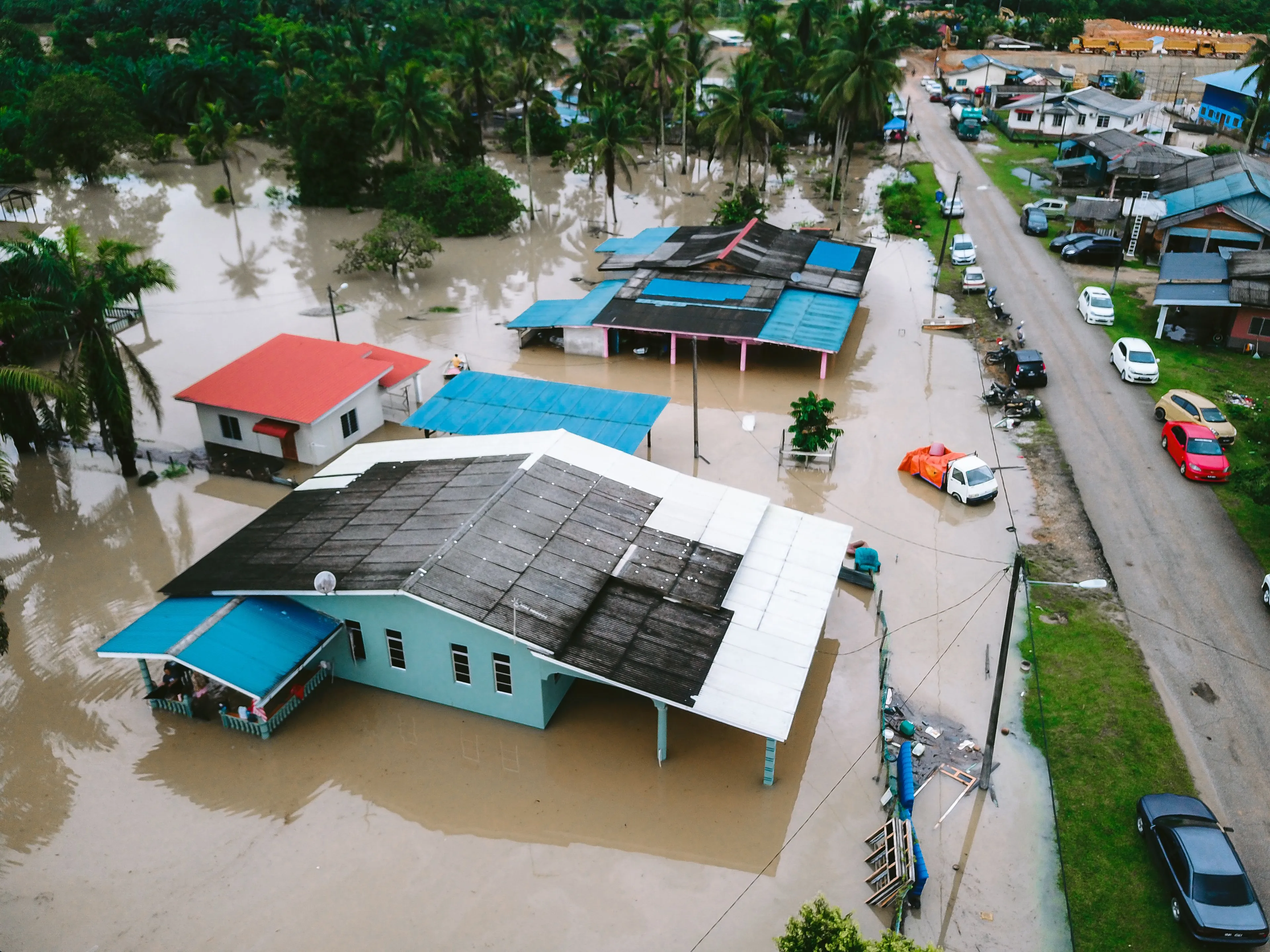 An aerial view of a village submerged in floodwaters, with houses partially visible and cars on a flooded road.