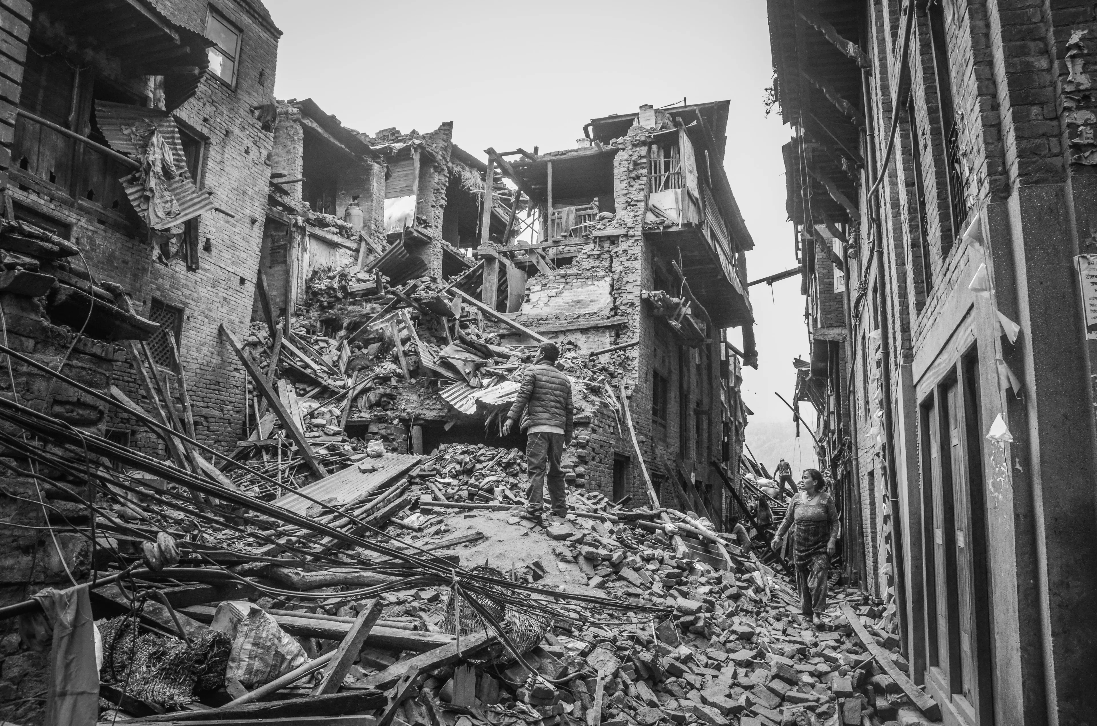 A black and white photo shows people walking through the rubble of buildings destroyed by an earthquake.