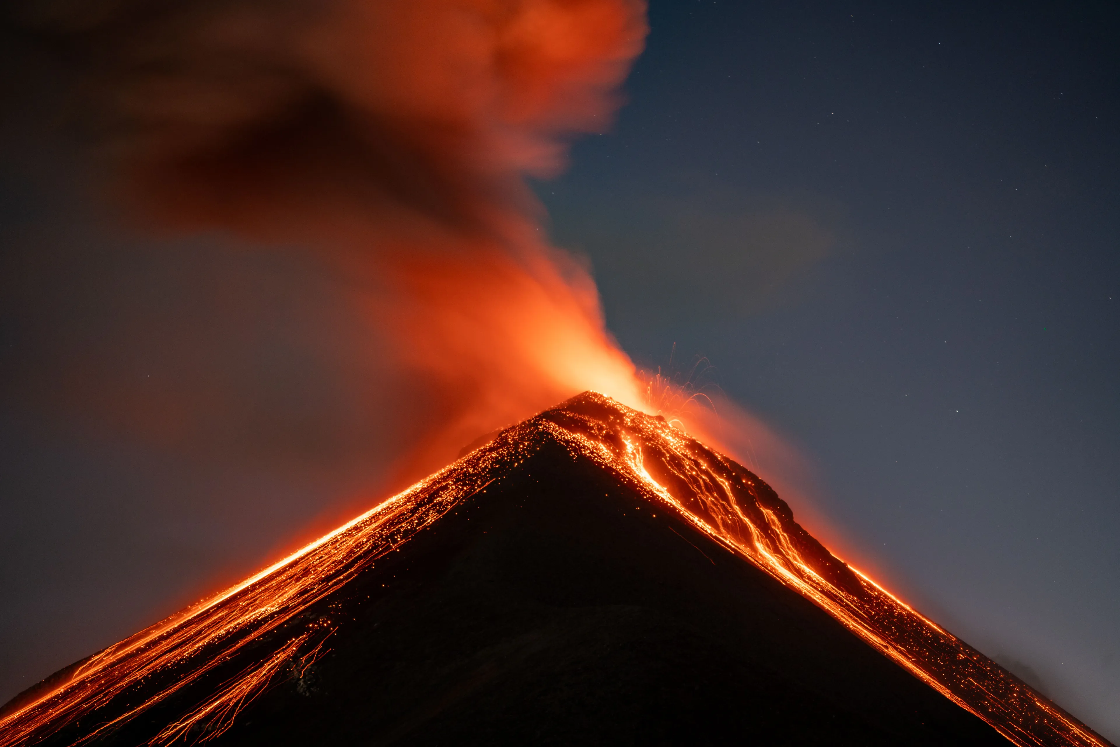 A dramatic night shot of a volcanic eruption with bright red lava flowing down the sides of the volcano.