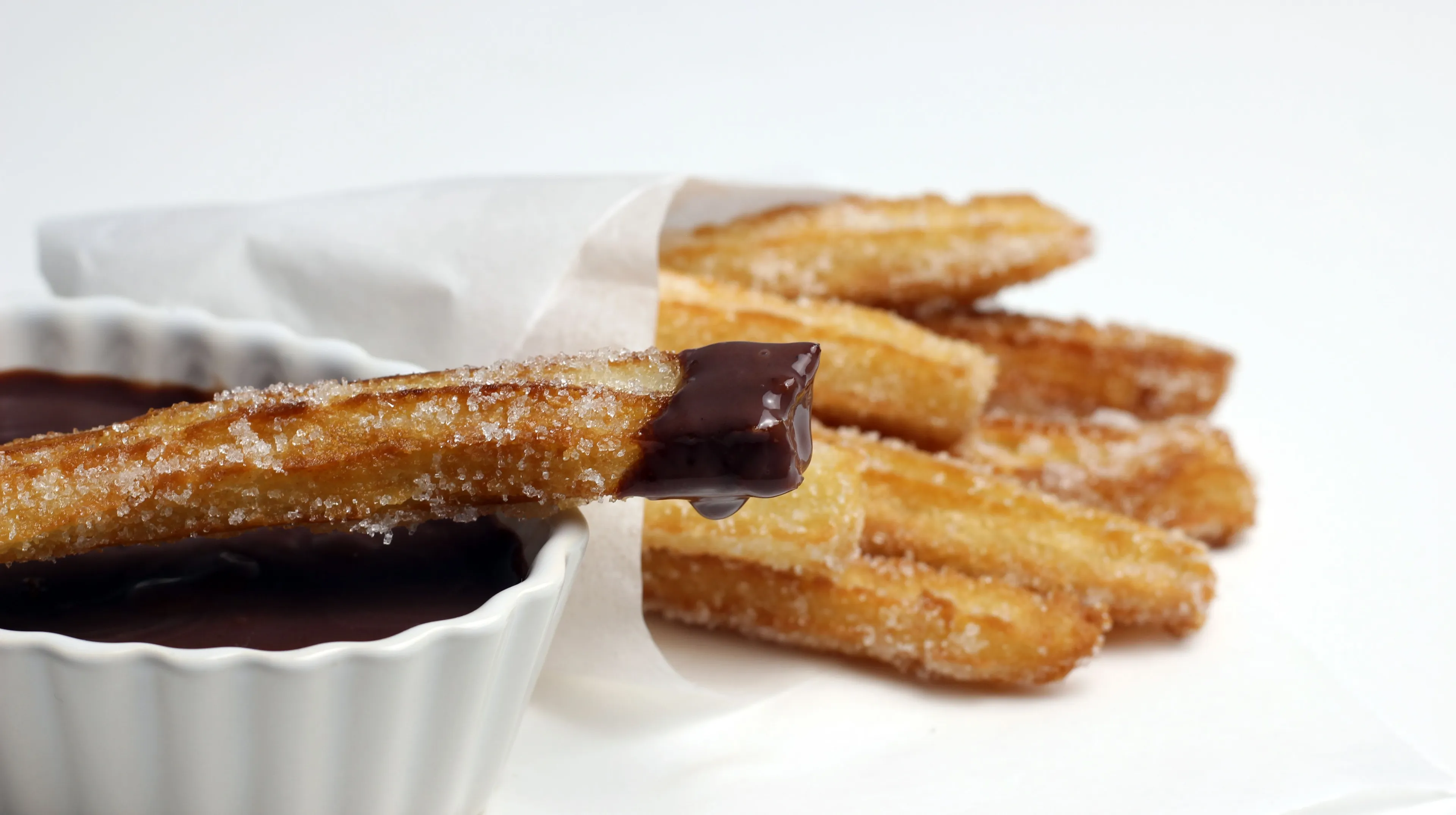 Crispy churros coated in sugar, one dipped in rich chocolate sauce, in a white paper bag.