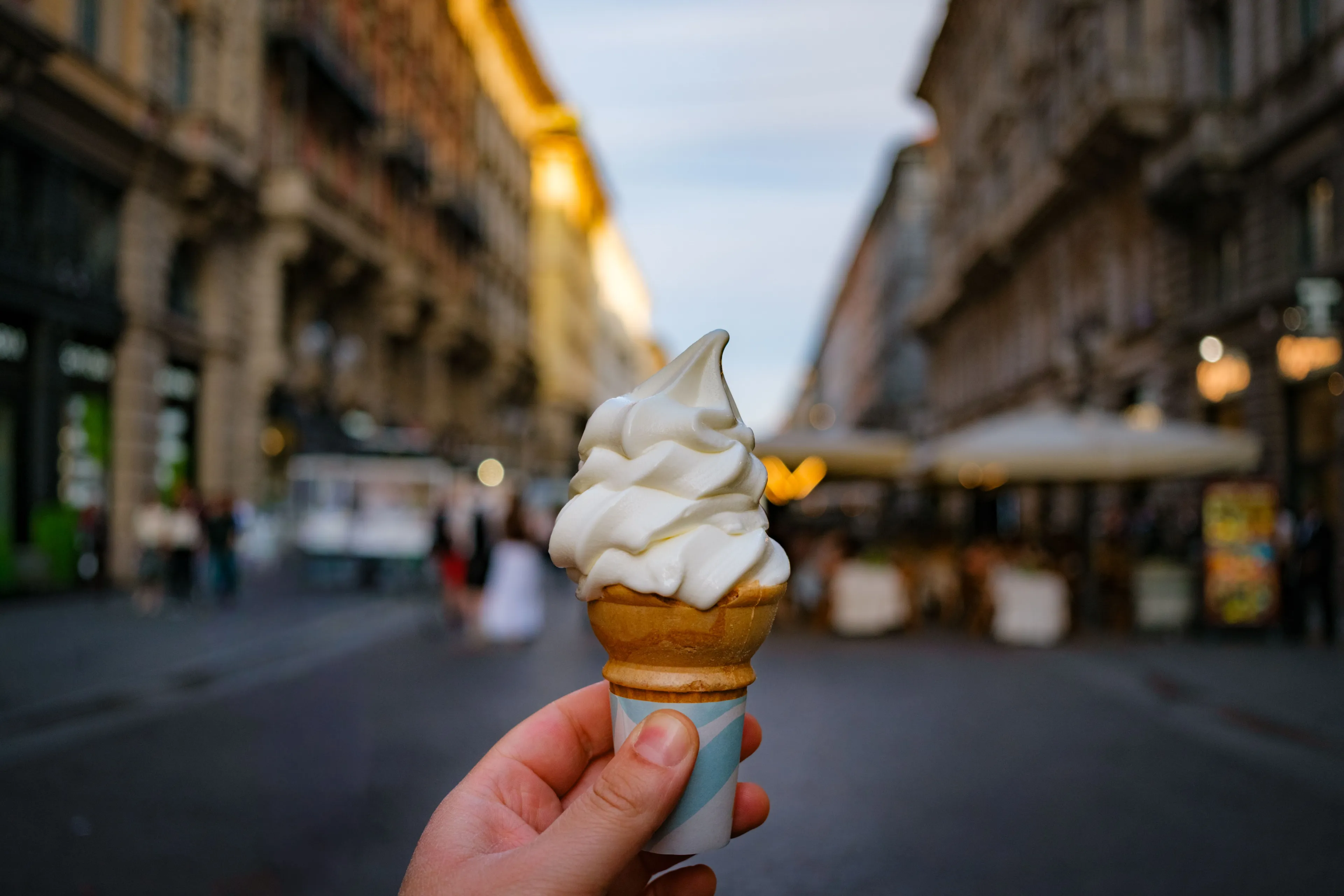 Hand holding a swirl of vanilla ice cream in a cone, walking down a city street.