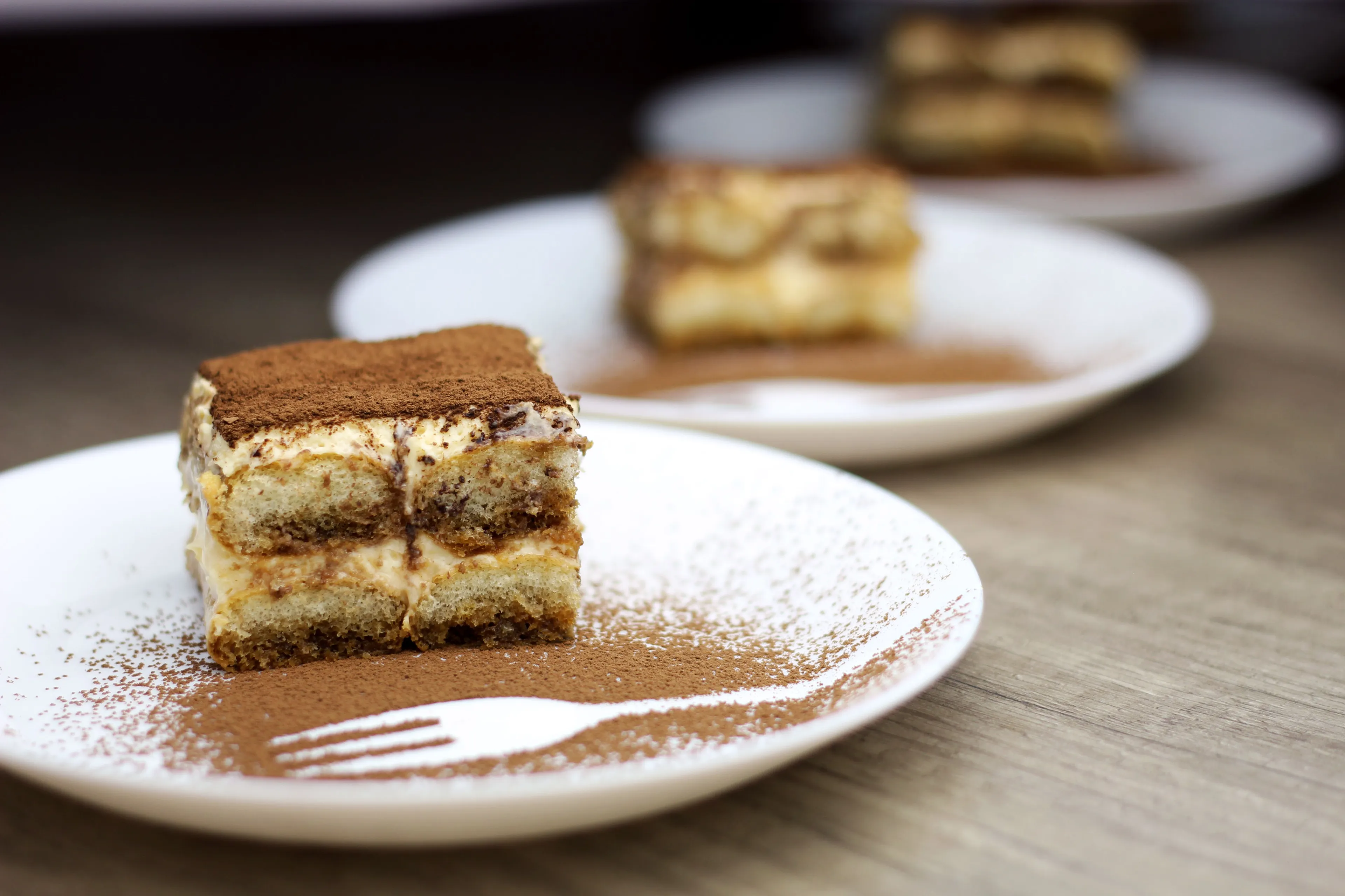 A slice of tiramisu dessert dusted with cocoa powder on a white plate with a fork design.