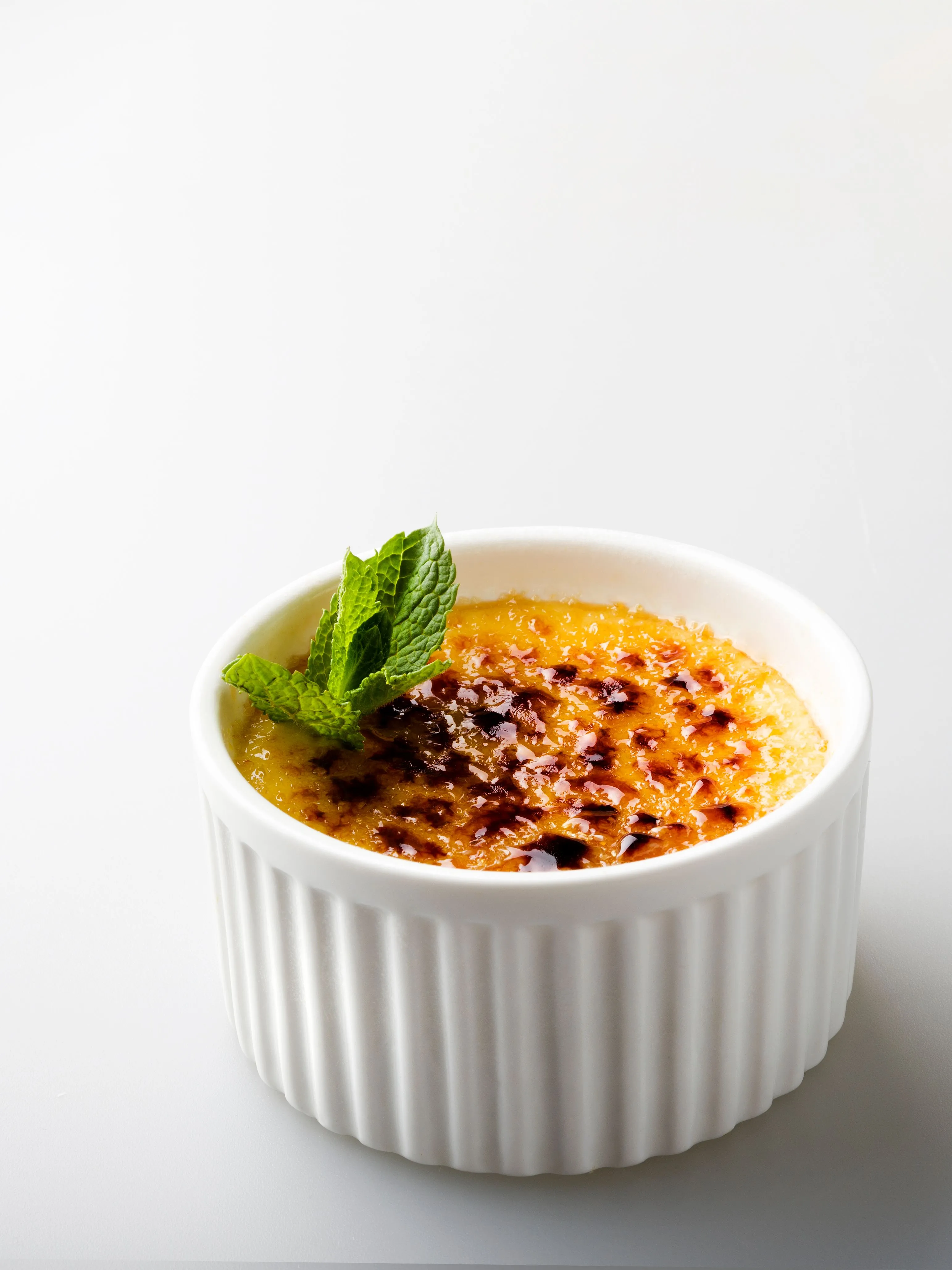 A close-up of a perfectly caramelized Crème brûlée in a white ramekin, garnished with a fresh mint leaf.