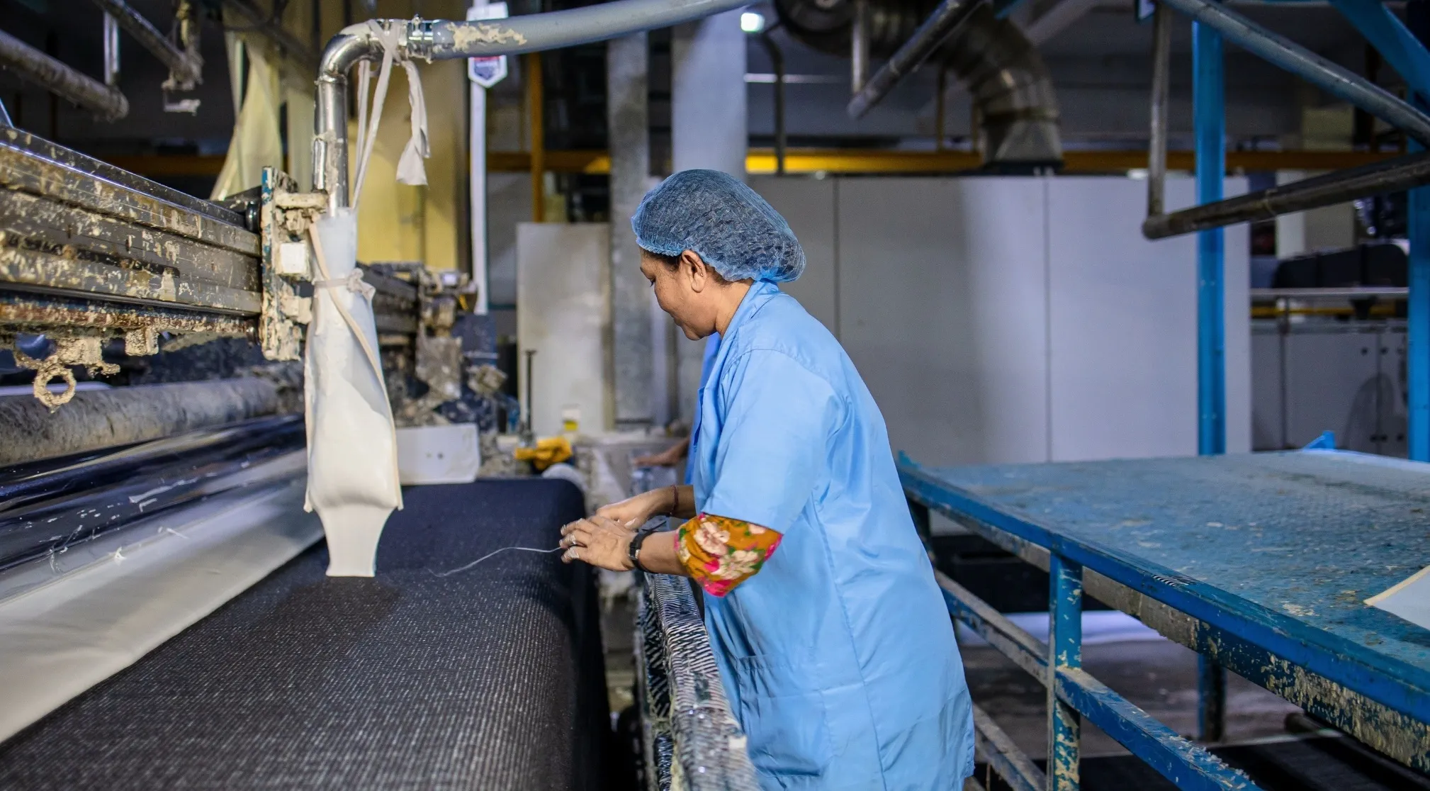 A textile manufacturer worker in a blue uniform and hairnet, tending to machinery producing fabric, showcasing the manufacturing process.