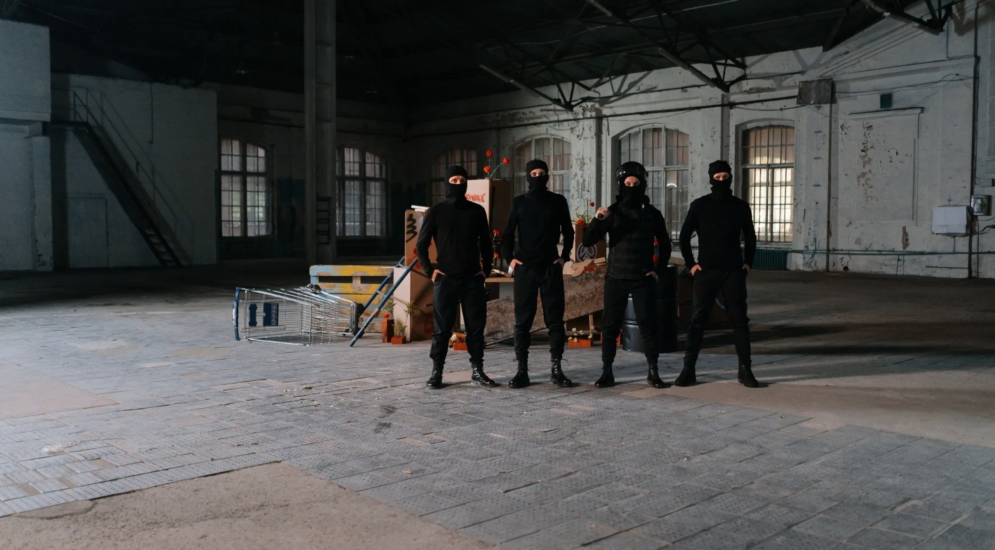 Four figures in black balaclavas and clothing in a dim, abandoned building, like WWI recruit test thieves committing depredations.