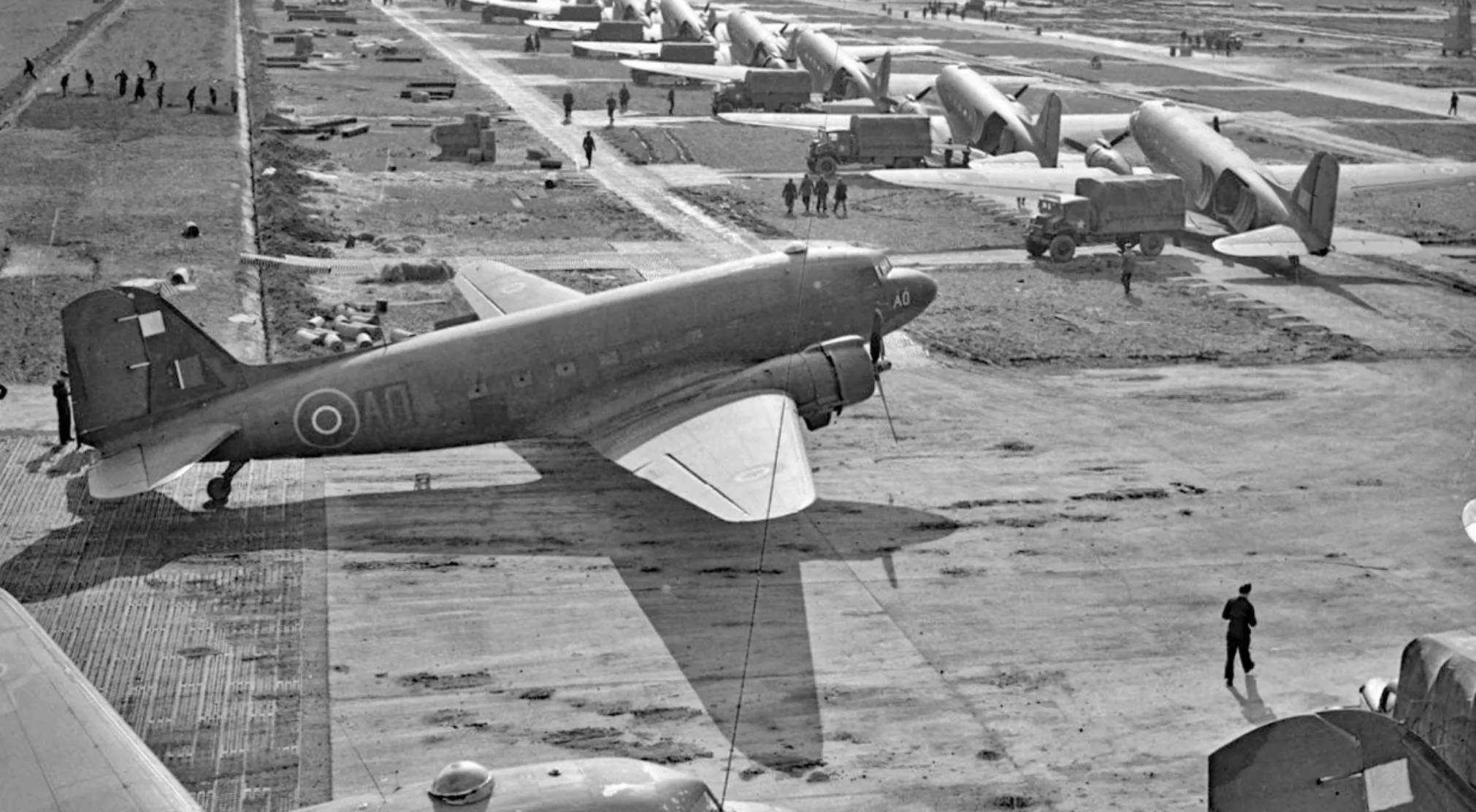 Black and white photo showing multiple World War II aircraft lined up on a rugged airfield with military personnel and trucks.