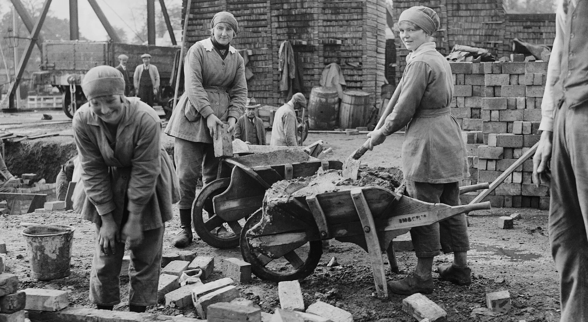 Historic black and white image of three women working at a construction site, loading bricks and mixing cement in wheelbarrows. Jeopardy Quiz.