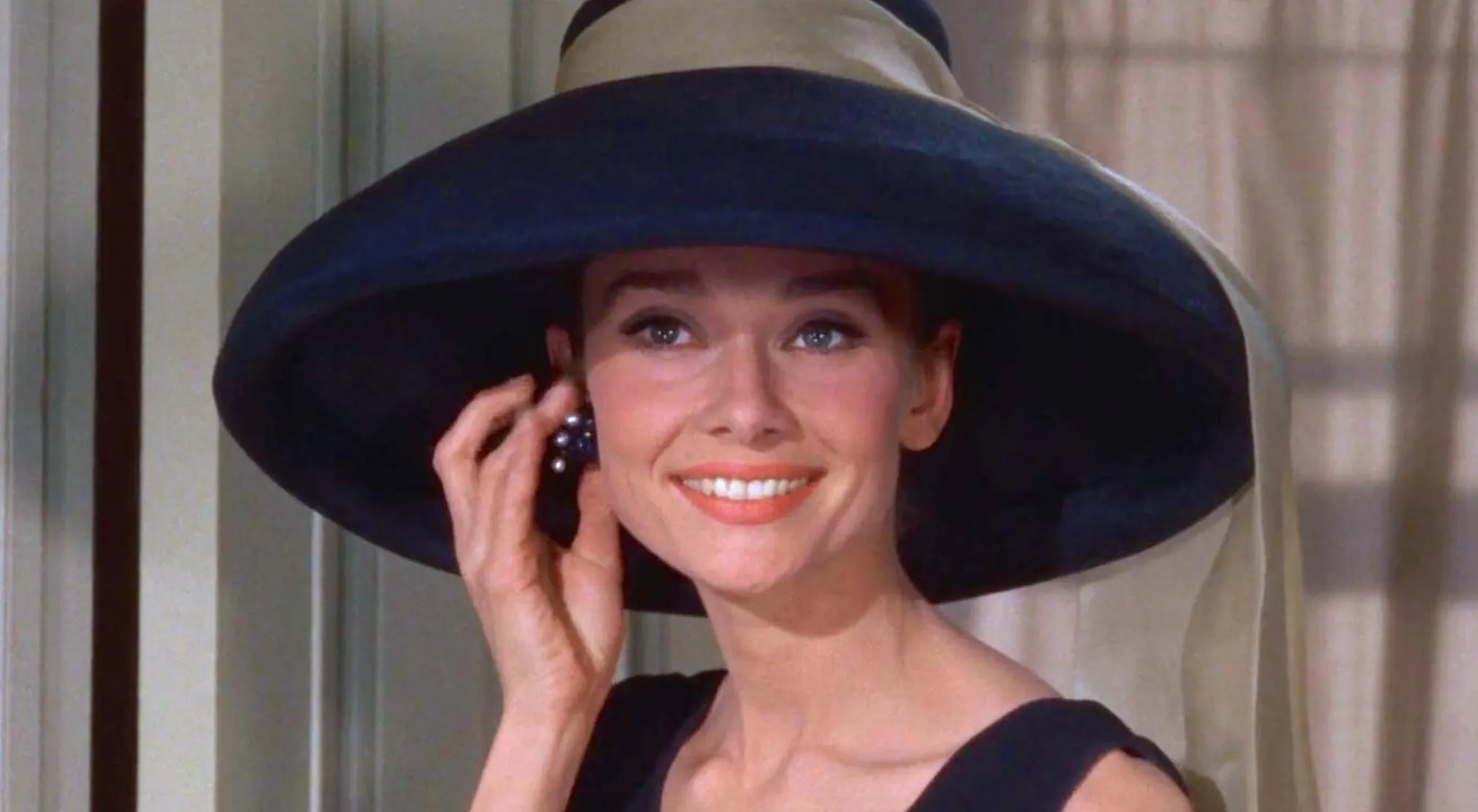 Audrey Hepburn from The Real Jeopardy Quiz Movies, wearing a large hat and smiling, adjusting her earring.