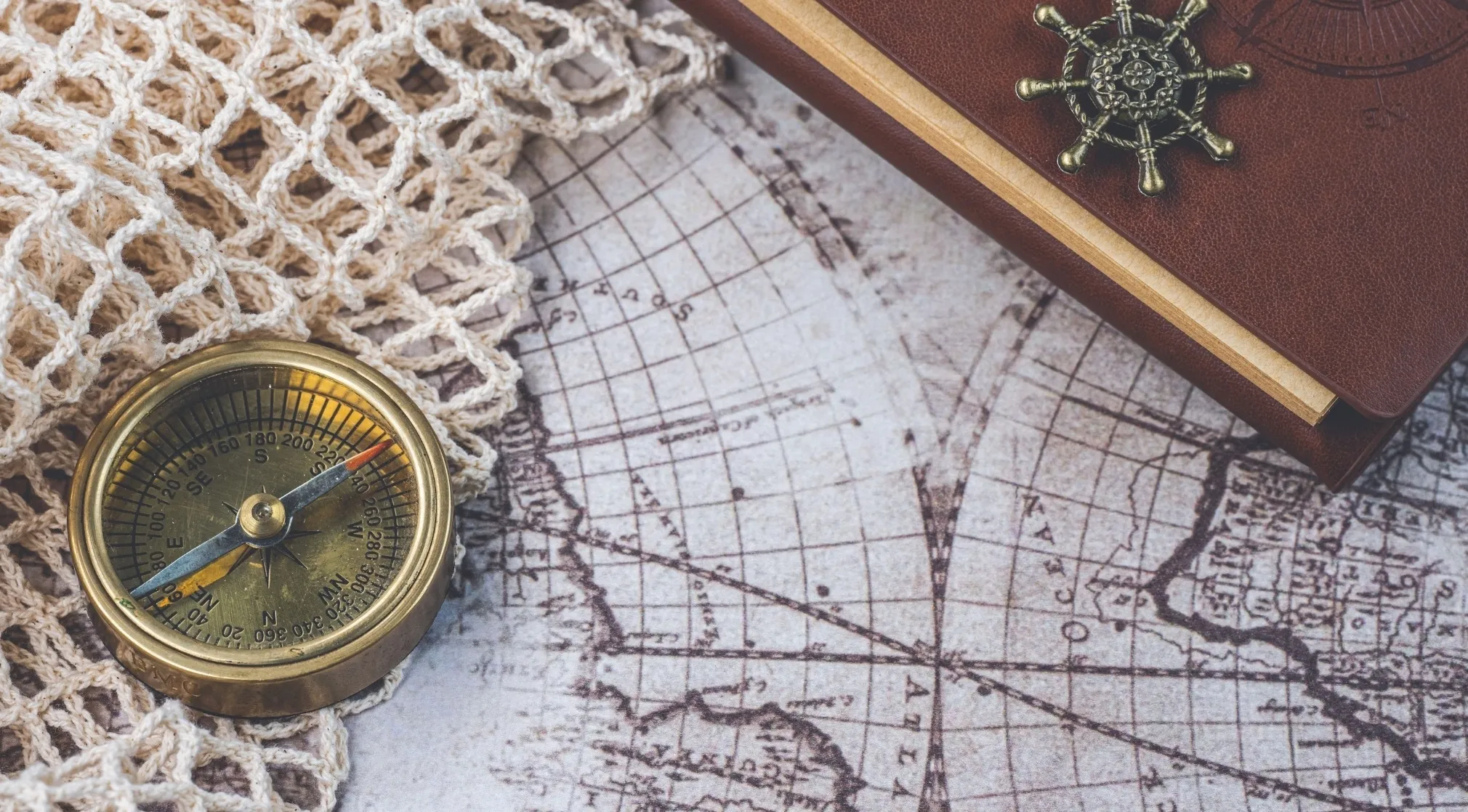 Vintage brass compass on a map, with a brown journal and ship wheel charm. Exploring remote oceans.