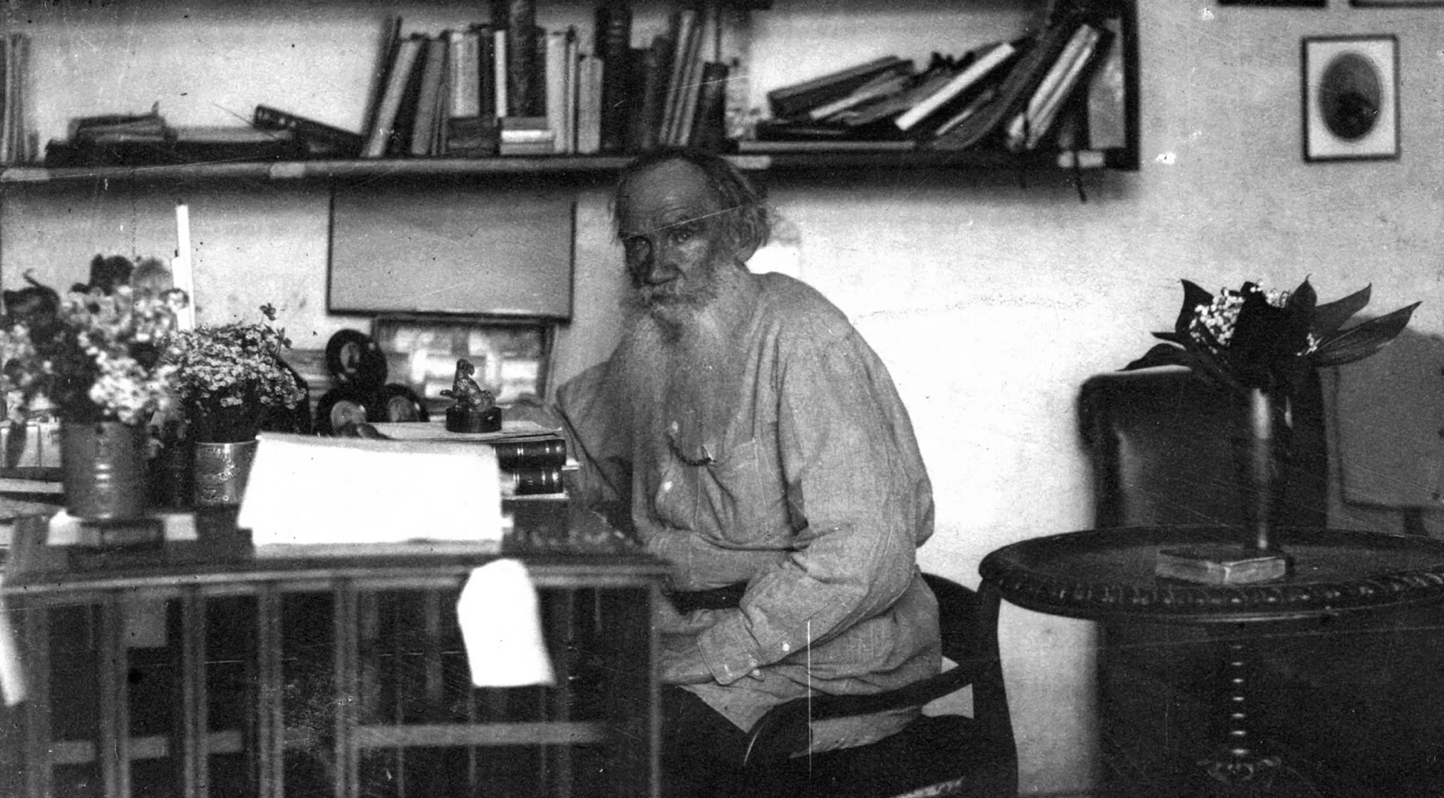 Black and white photo of a bearded old man, possibly Tolstoy, in a room with a bookshelf and flowers. Jeopardy Quiz Literature.