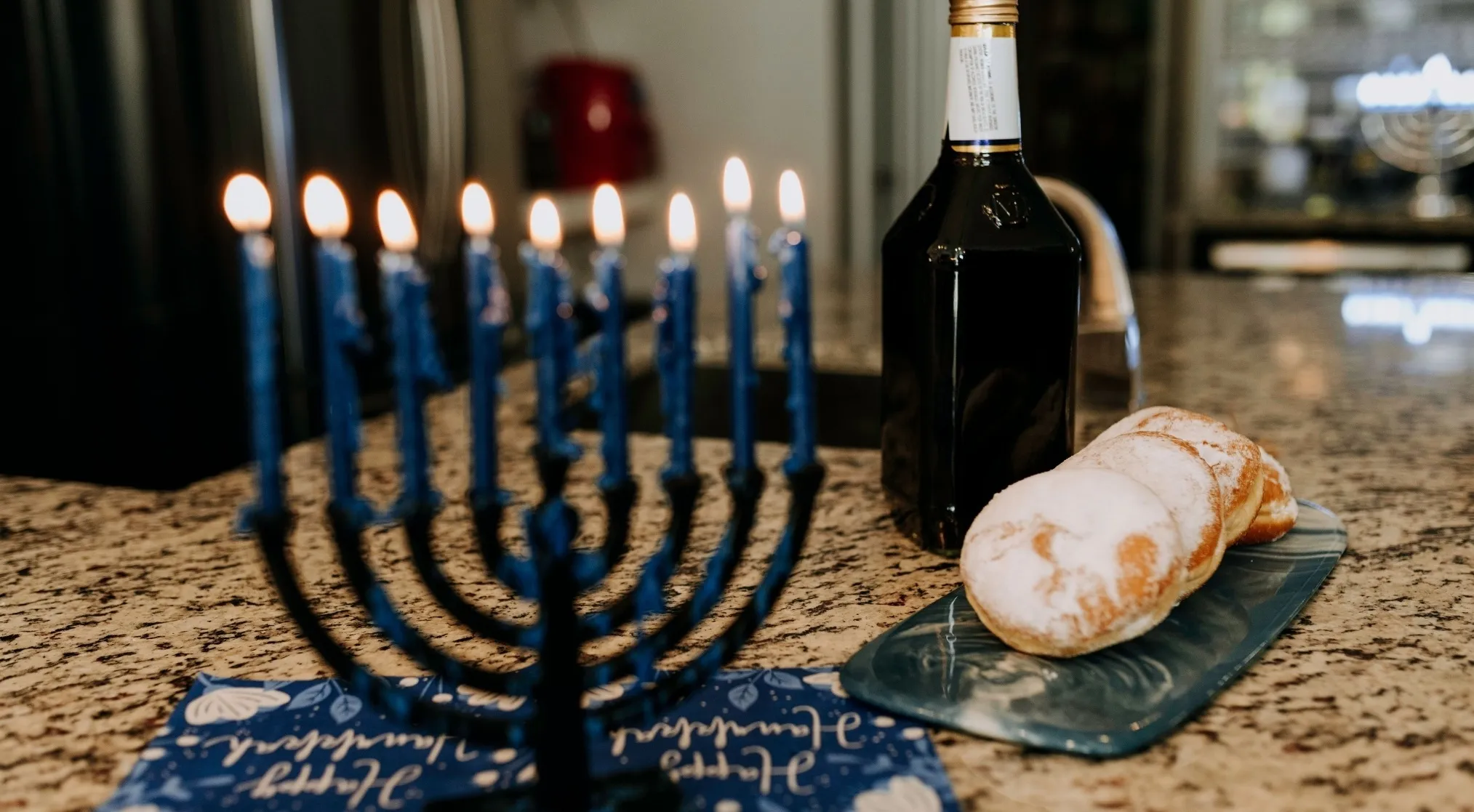 A menorah with lit blue candles, a bottle, and powdered sugar donuts on a counter. Real Jeopardy Quiz Celebrities.