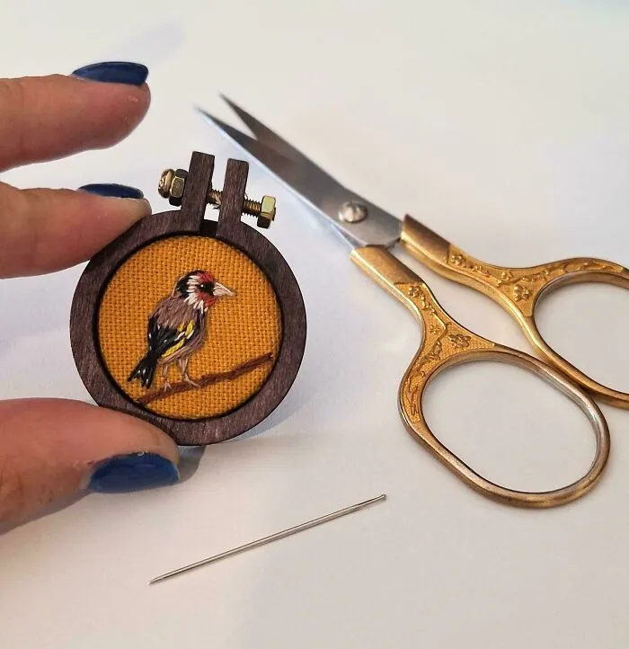A tiny hand holding a miniature bird embroidery project next to scissors