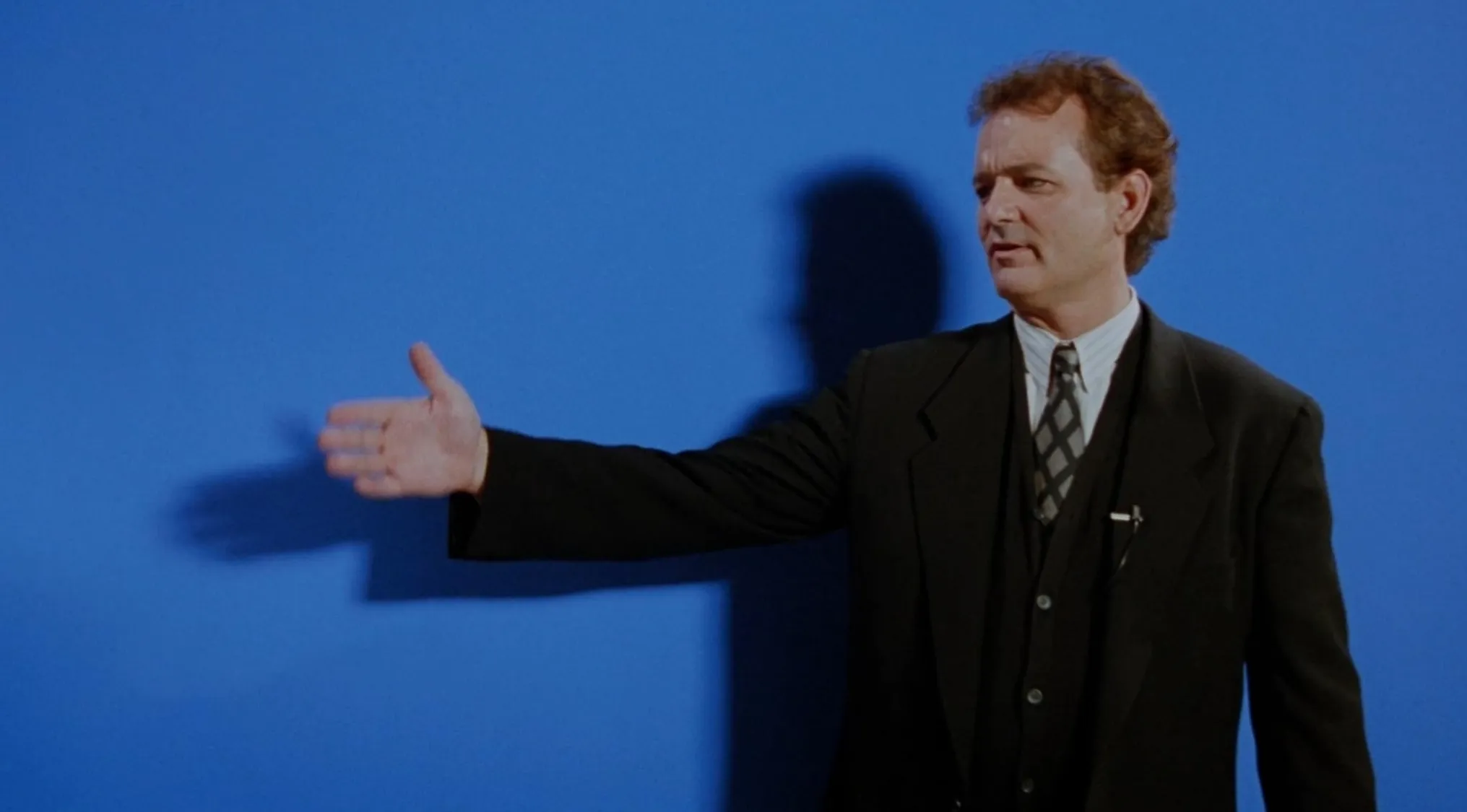 A weatherman in a black suit gestures on a blue screen in an opening movie scene.