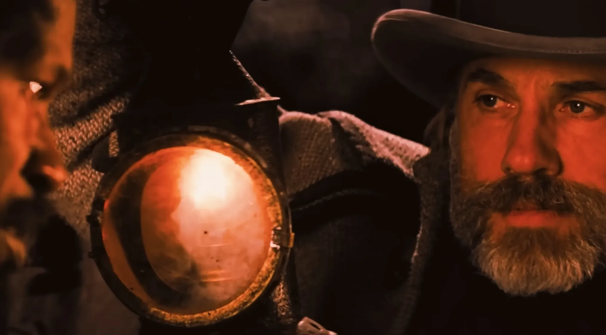 A man with a beard and hat shines a lantern on another person in a dark opening movie scene.