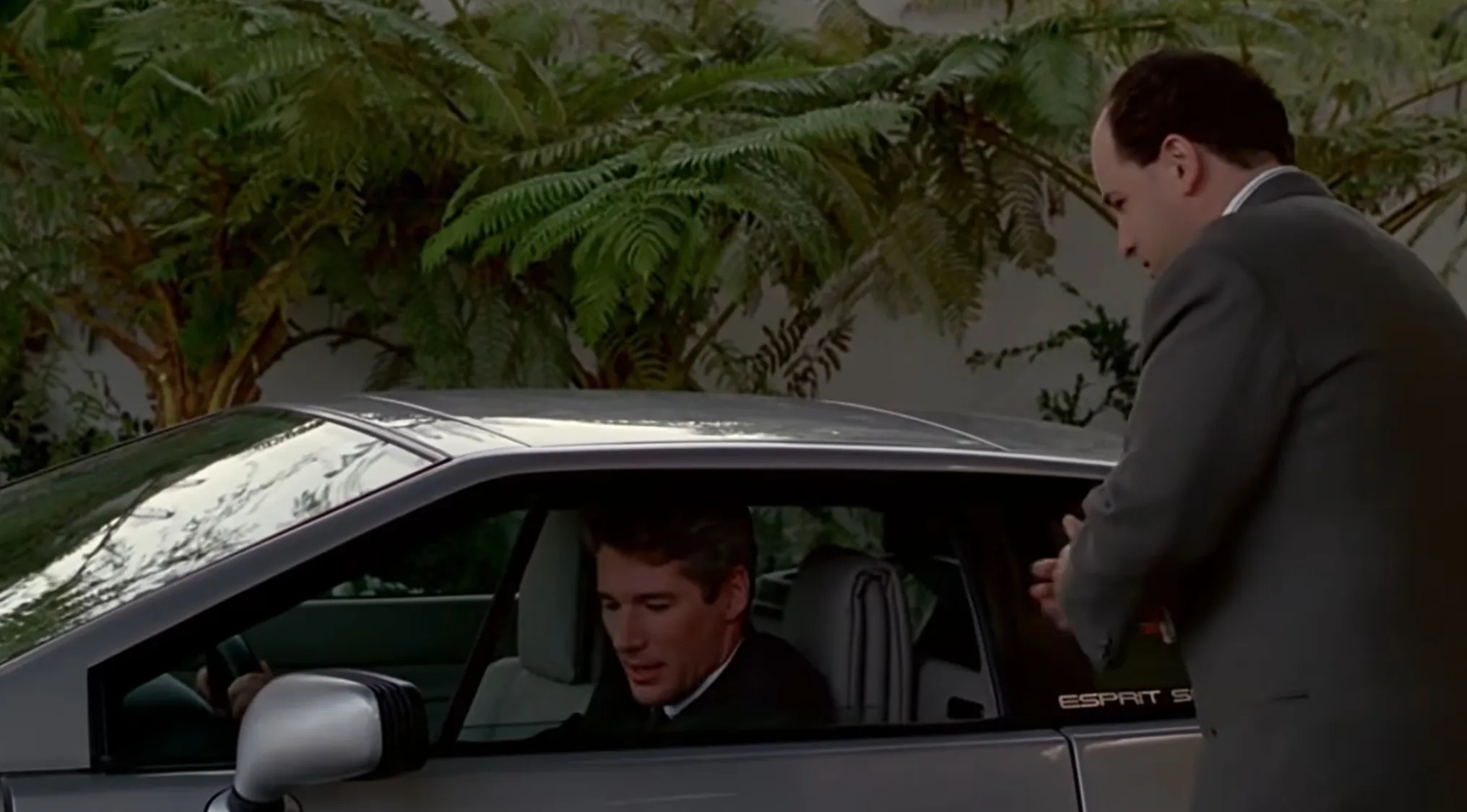 A man in a suit in a car, looking down, while another man stands outside the car in an opening movie scene.