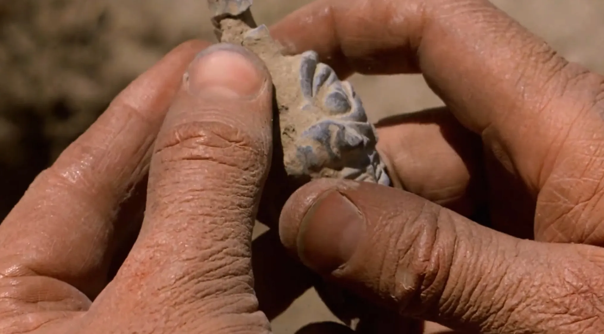 A pair of hands gently holding an ancient, dirty amulet with intricate carvings, found at an archaeological site.