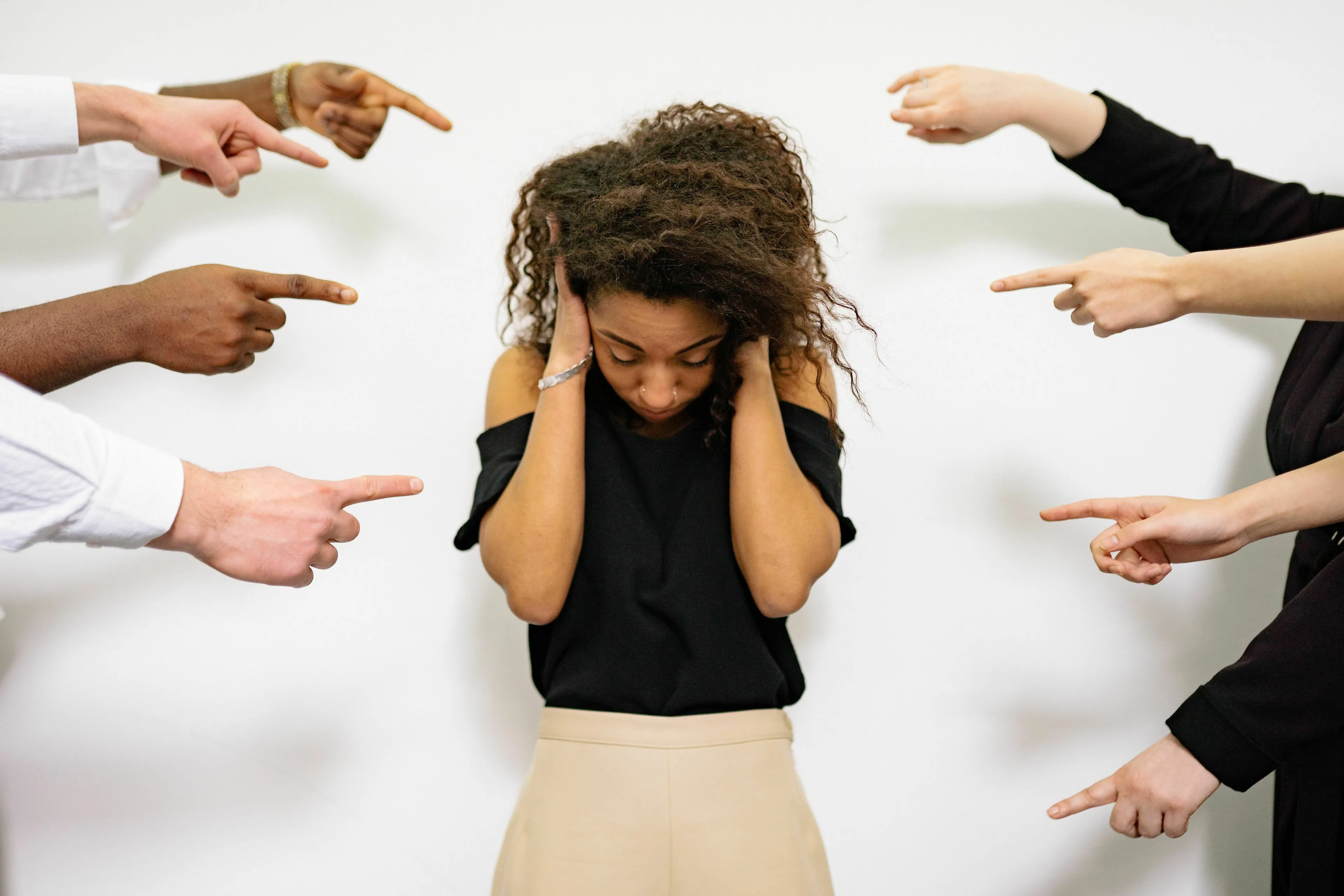 A young woman with curly hair covering her ears while multiple hands point at her, illustrating feeling misunderstood.