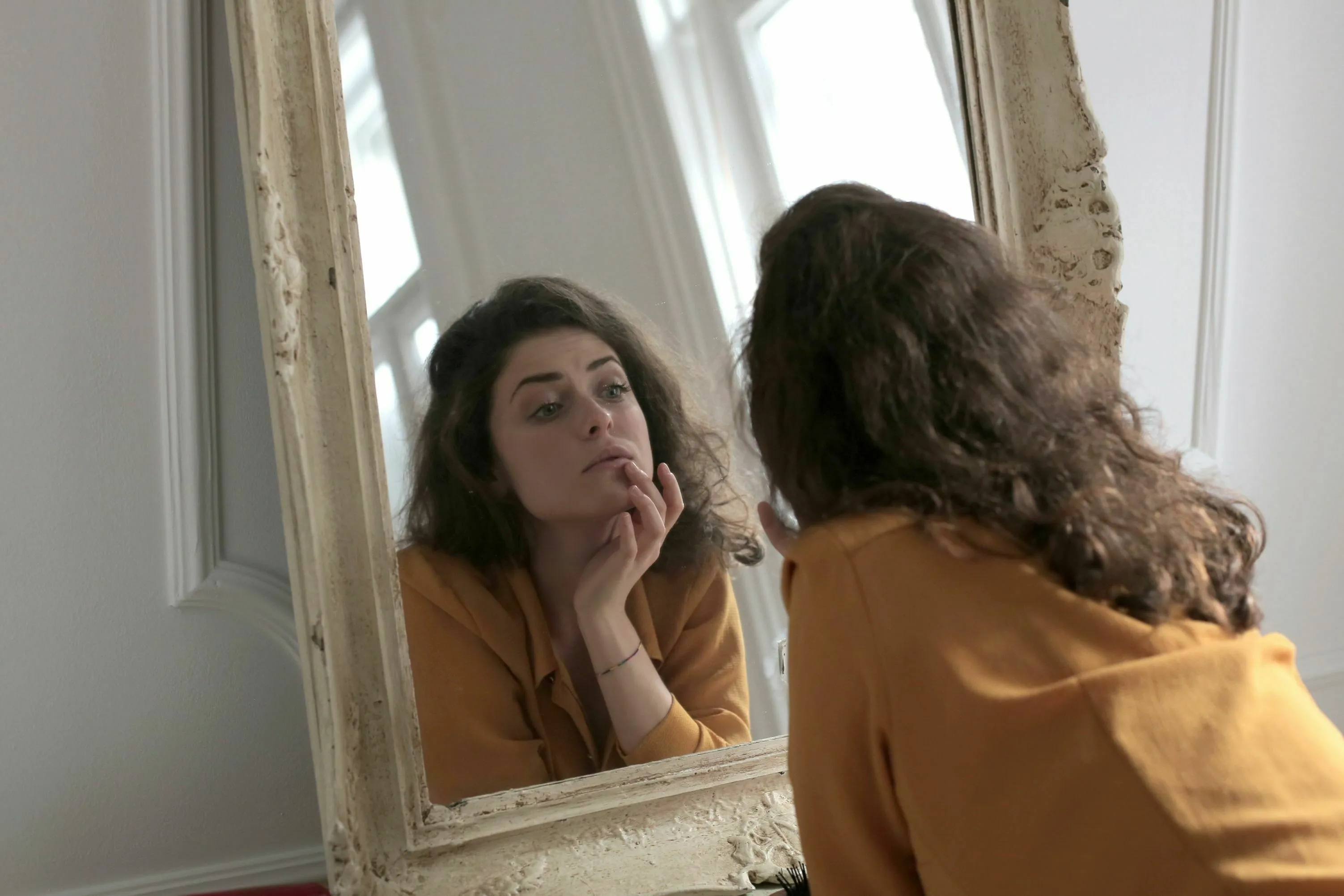 A woman with curly hair looking intently at her reflection in a large, ornate mirror, seeking validation for her feelings.