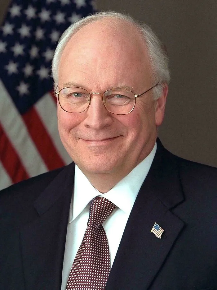 Close-up portrait of Dick Cheney smiling in a suit with an American flag, referencing timeline facts.