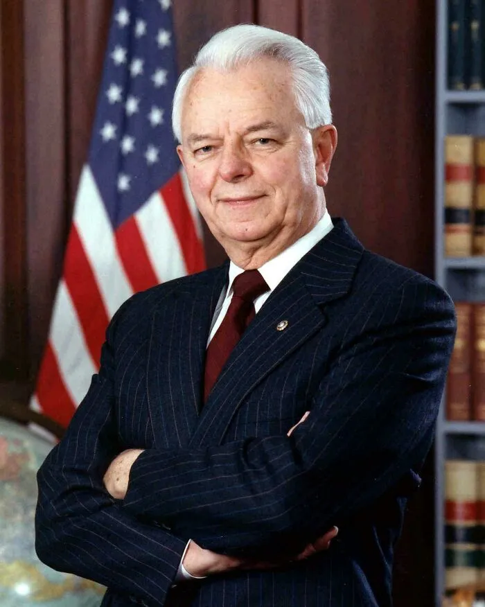 A distinguished older man in a suit, standing with the American flag in the background, offering historical facts.