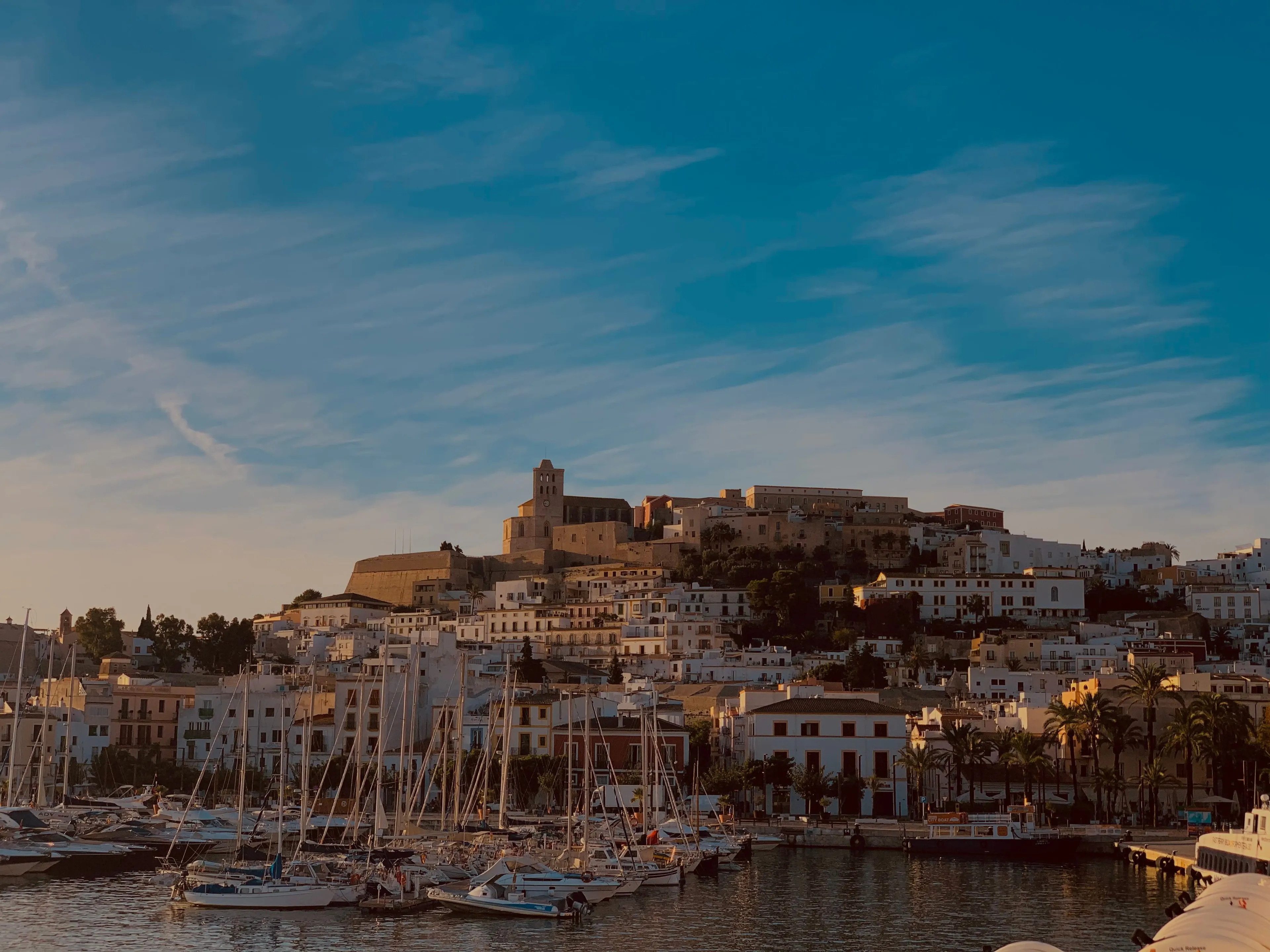 A picturesque view of Ibiza town from the harbor, with boats and whitewashed buildings under a blue sky, an ideal vacation spot.