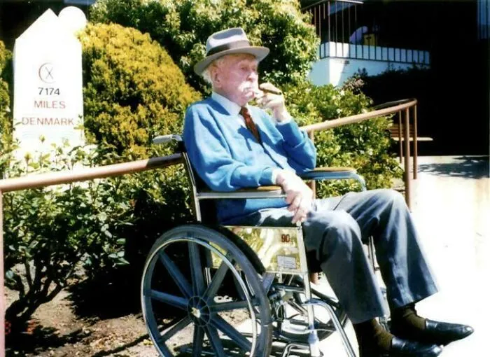 An elderly man in a wheelchair smoking, next to a sign showing historical timeline facts.